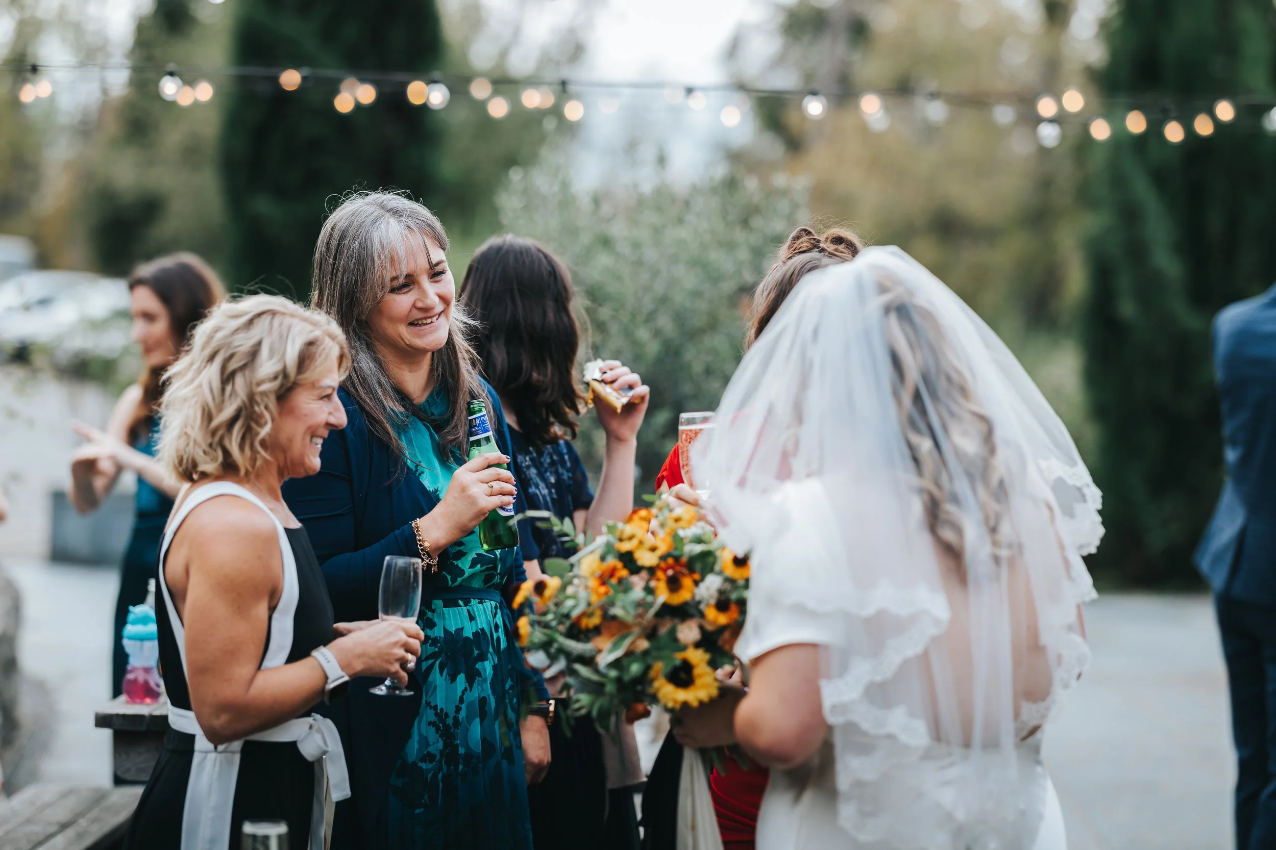 Group of women at outdoor wedding reception, smiling, holding drinks, with a bride in a white dress and veil holding a bouquet of sunflowers and smiling