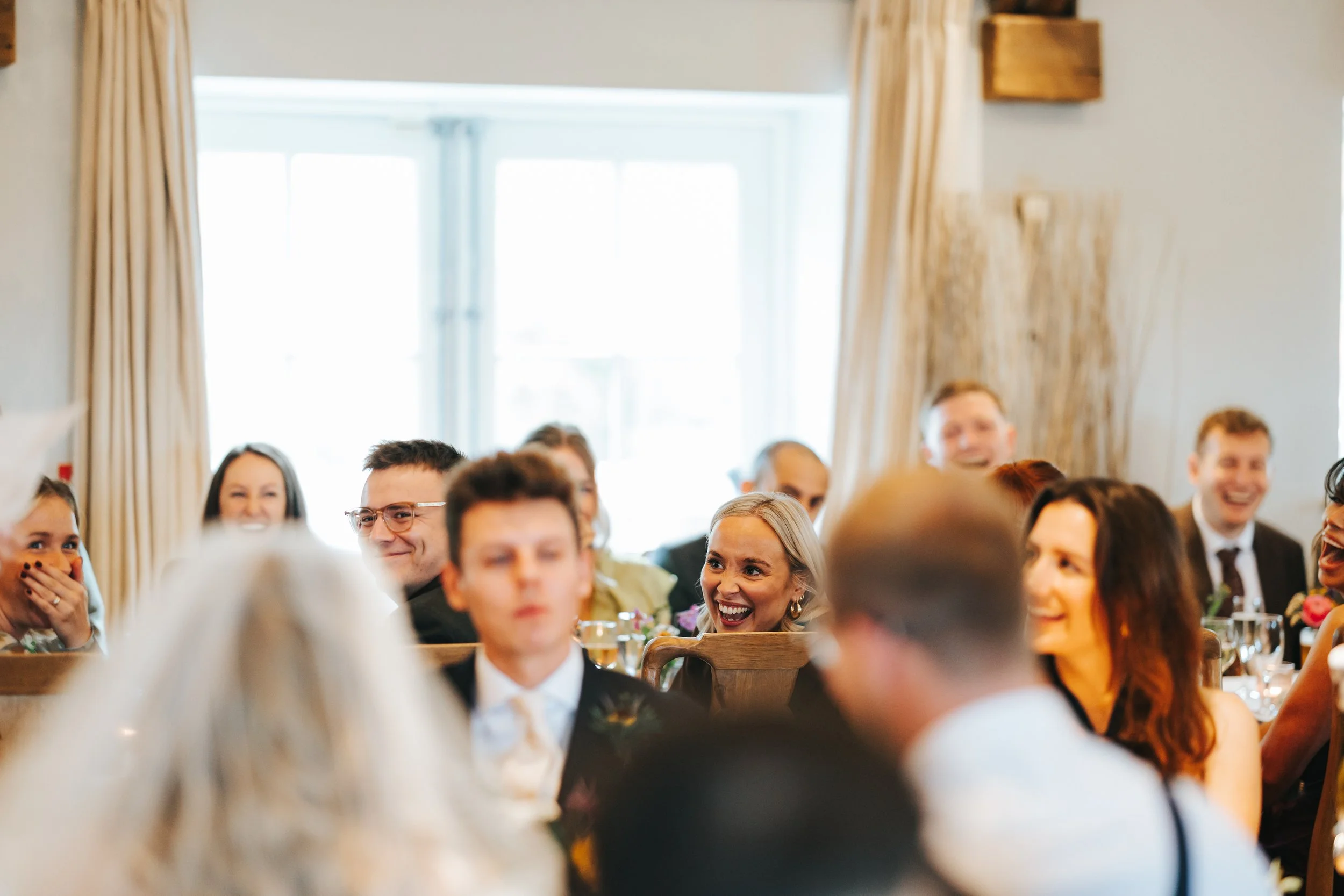 Group of people sitting at a table, smiling and laughing during a celebration in a bright room with large windows and beige curtains.