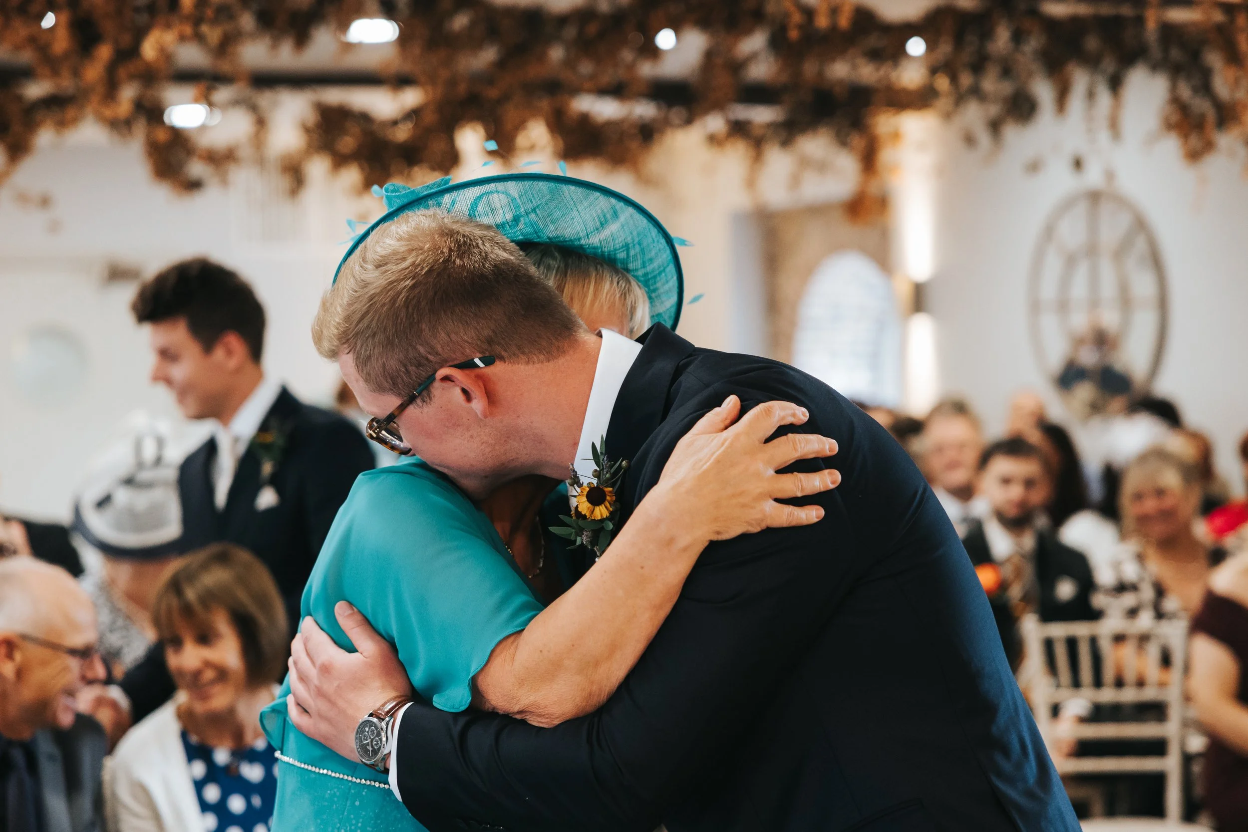 A man in a suit and glasses hugging an older woman wearing a turquoise dress and a hat with blue accents at a social event, with guests in formal attire seated in the background.