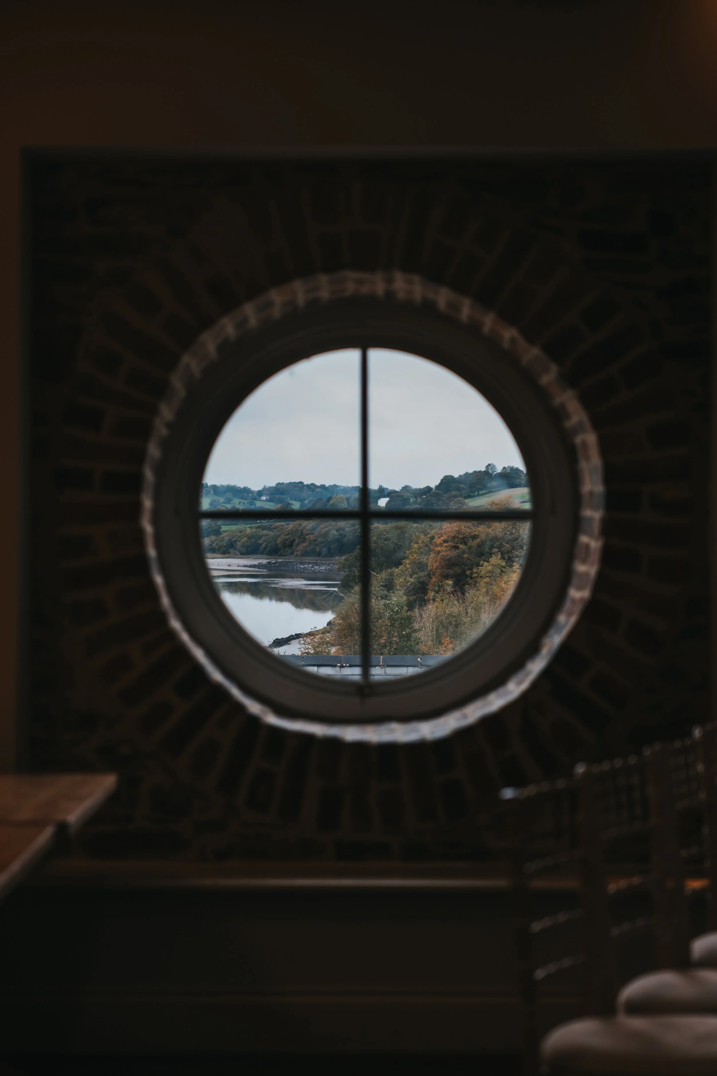 Round window with crossbars, showing a river, trees, and hills outside, inside a room with brick framing around the window.