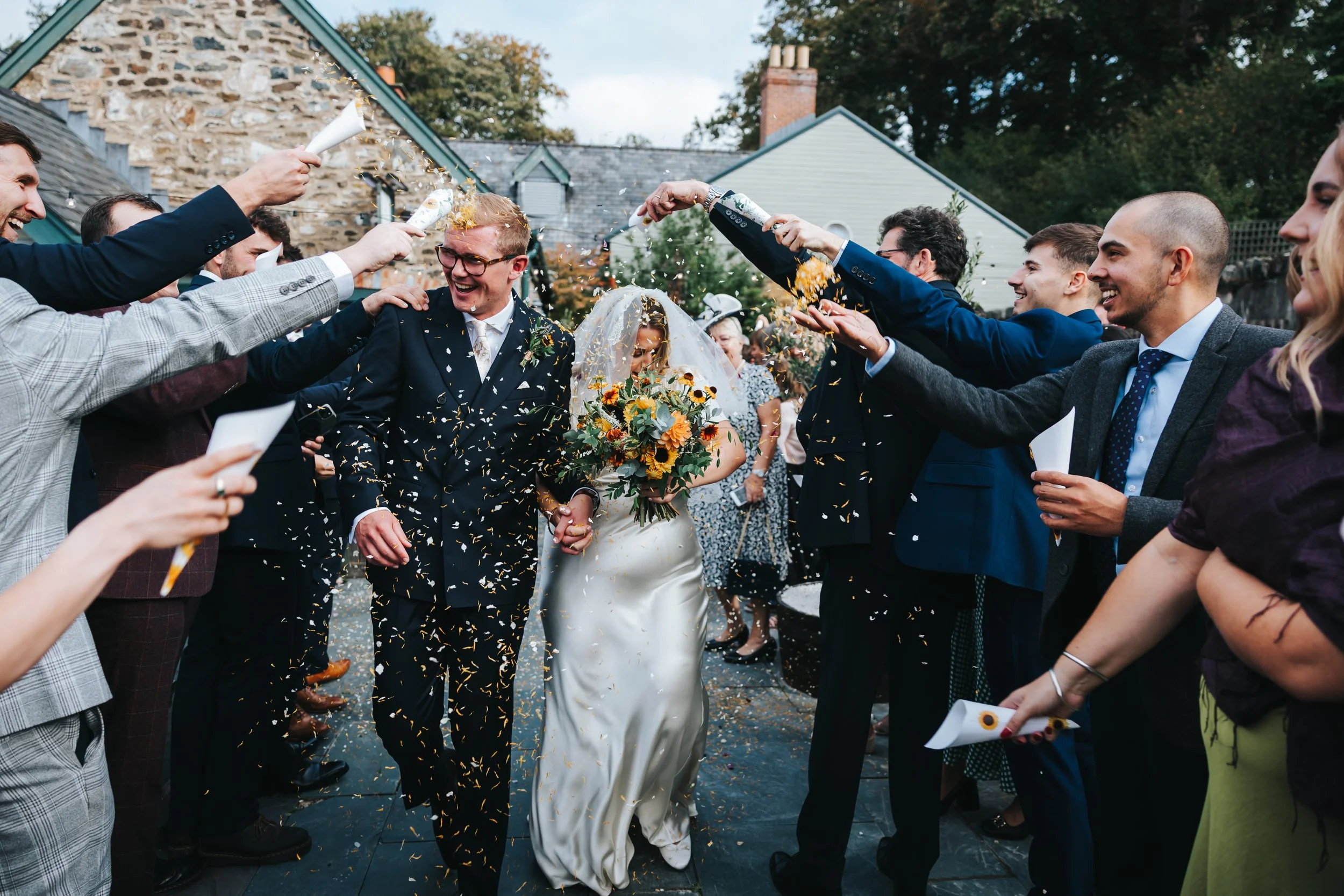 Bride and groom walking through a confetti throw at their wedding surrounded by guests outdoors.