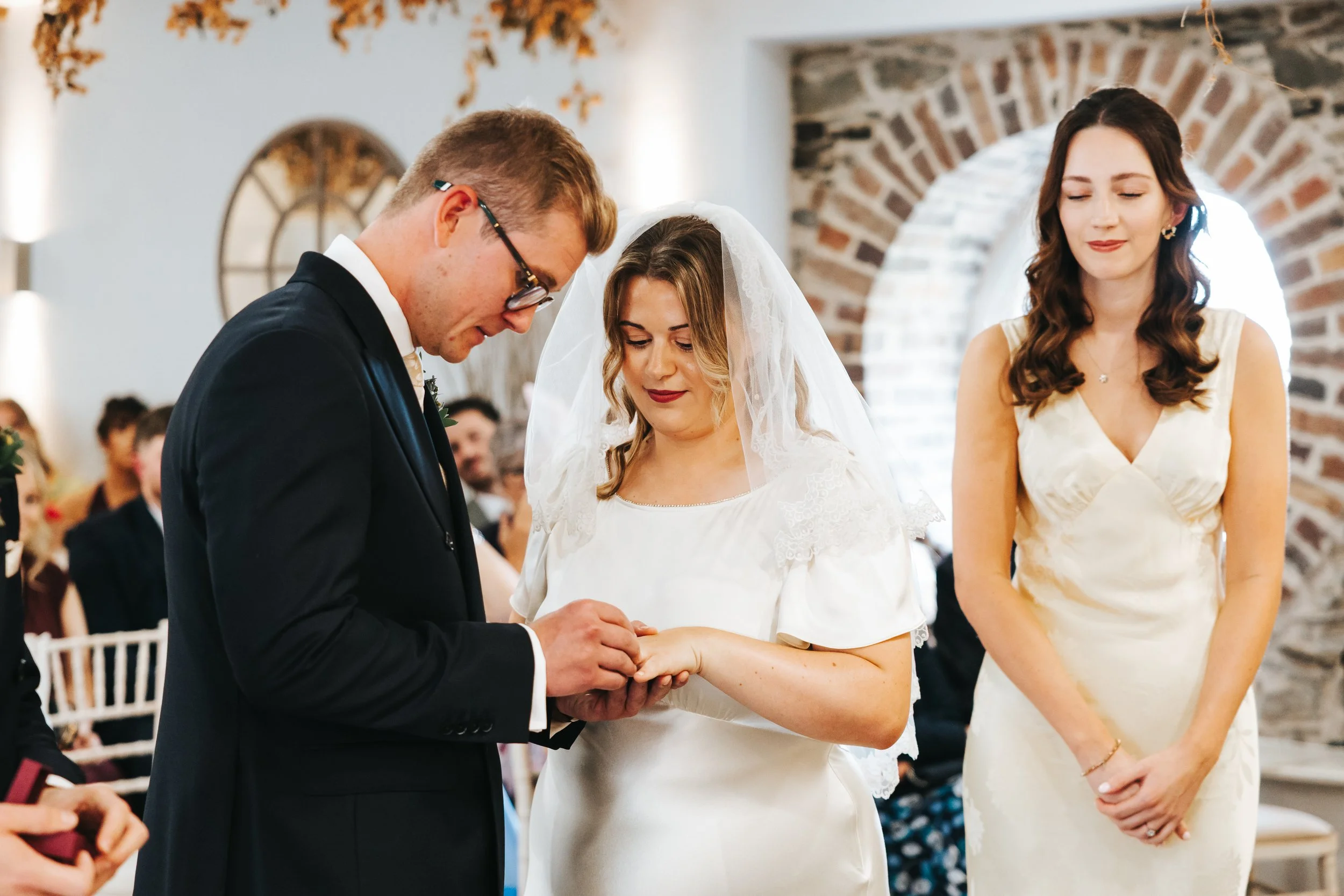 A wedding ceremony where a bride and groom are exchanging rings, with a woman in a cream dress standing beside them.