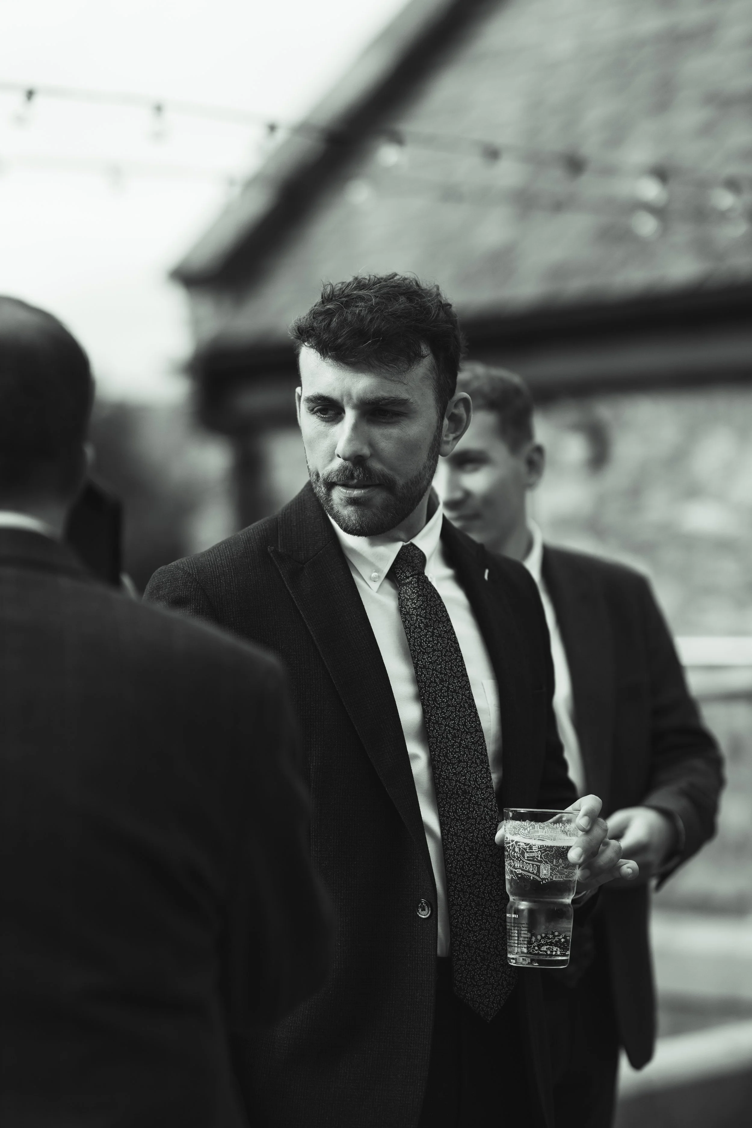 Black and white photo of three men in suits having a conversation, one holding a glass of water, outdoors near a building.
