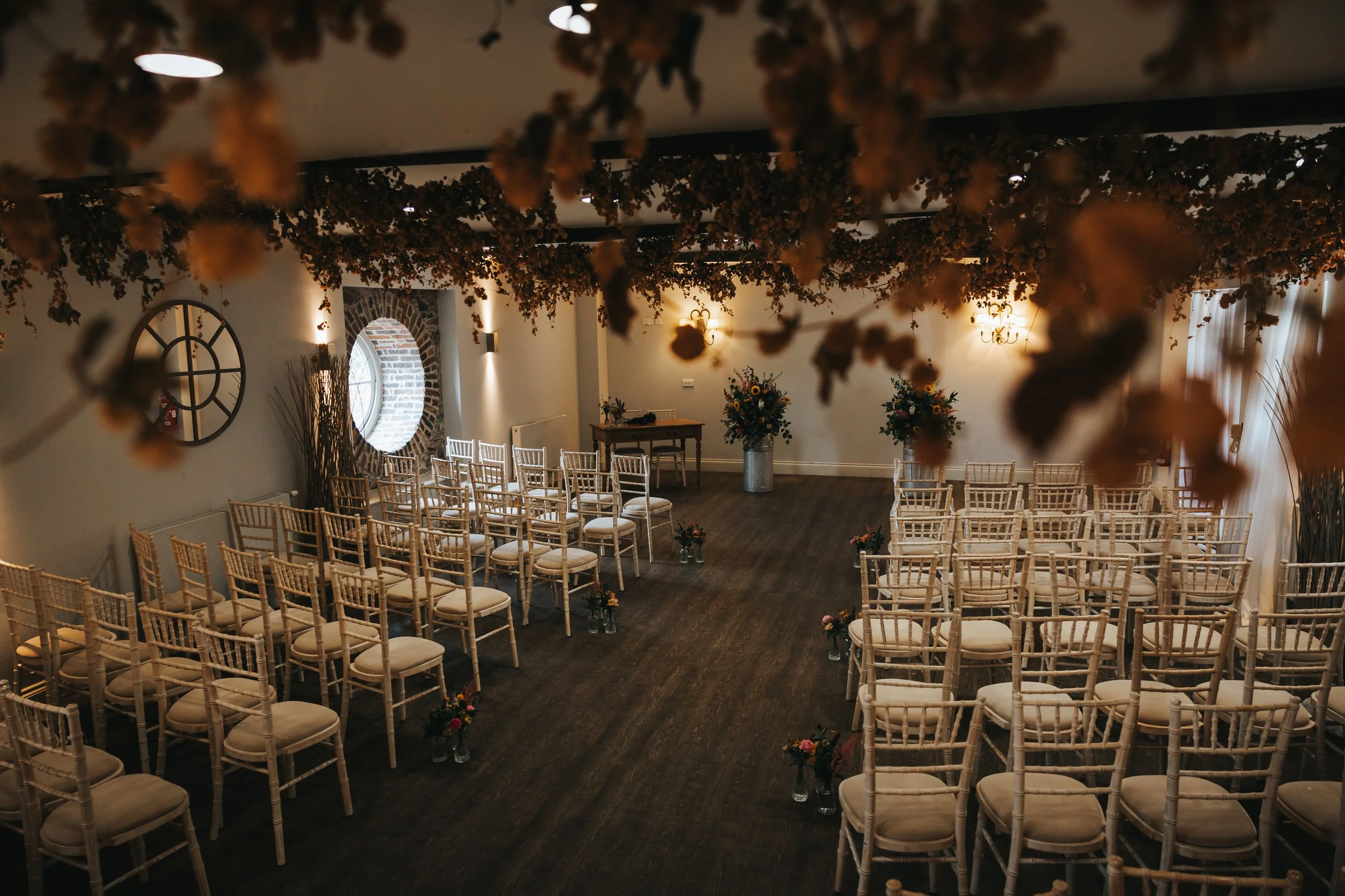 Empty wedding ceremony room with rows of white chairs, floral arrangements, and overhead decorative dried flowers.