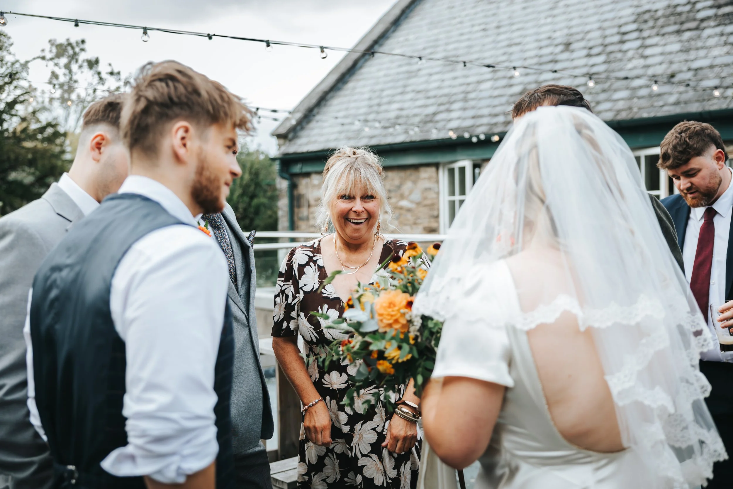 A group of people at a wedding reception outdoors, including a woman holding a bouquet and a bride with a veil, smiling and talking to guests.