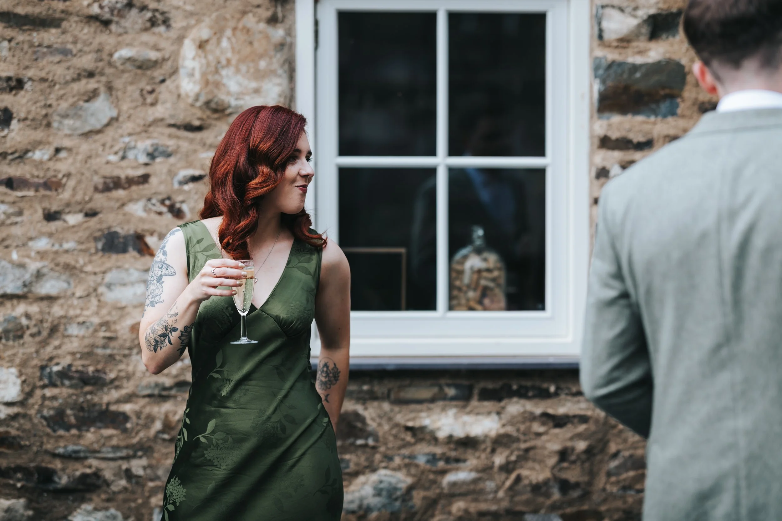 A woman with red hair and tattoos on her arms, wearing a green dress, holding a glass of champagne, standing outside a stone building with a window, looking at a man in a gray suit.