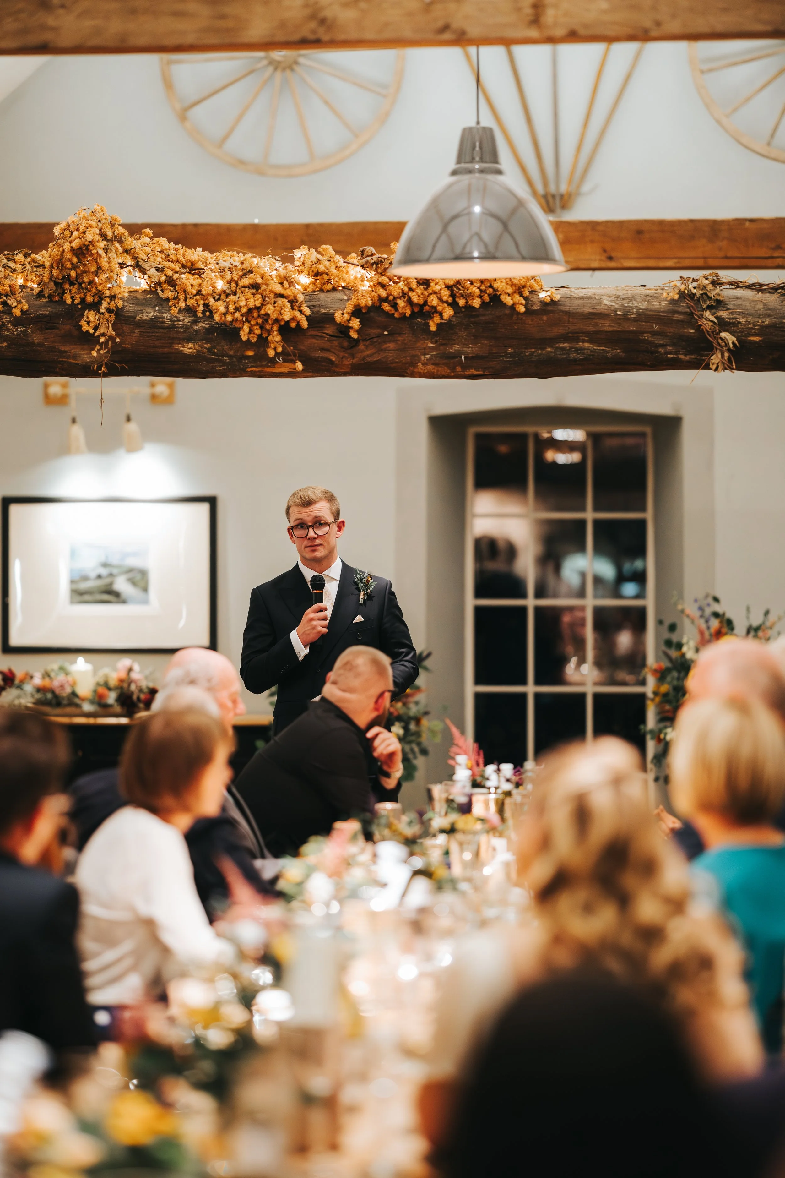 A man in a suit giving a speech at a wedding reception, with guests seated at a long table decorated with flowers and candles, inside a rustic venue with wooden beams and wheel decorations on the wall.