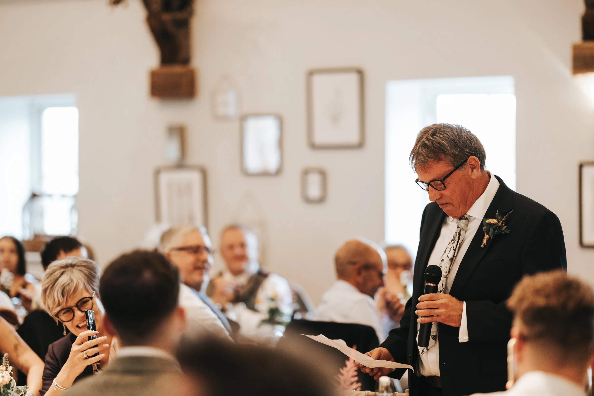 A man in a black suit and glasses speaking into a microphone at a wedding reception, with guests sitting at tables in the background.