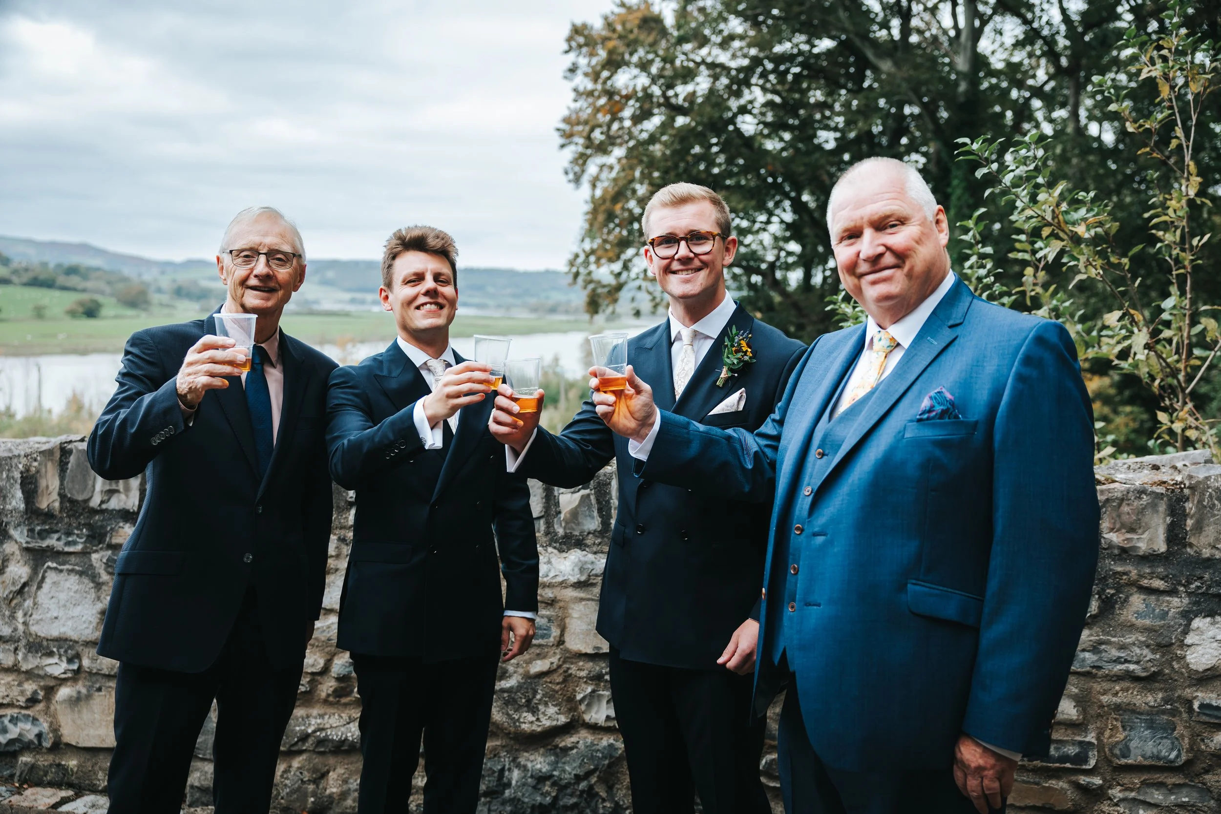 Four men in suits holding drinks and smiling outdoors at a celebration.