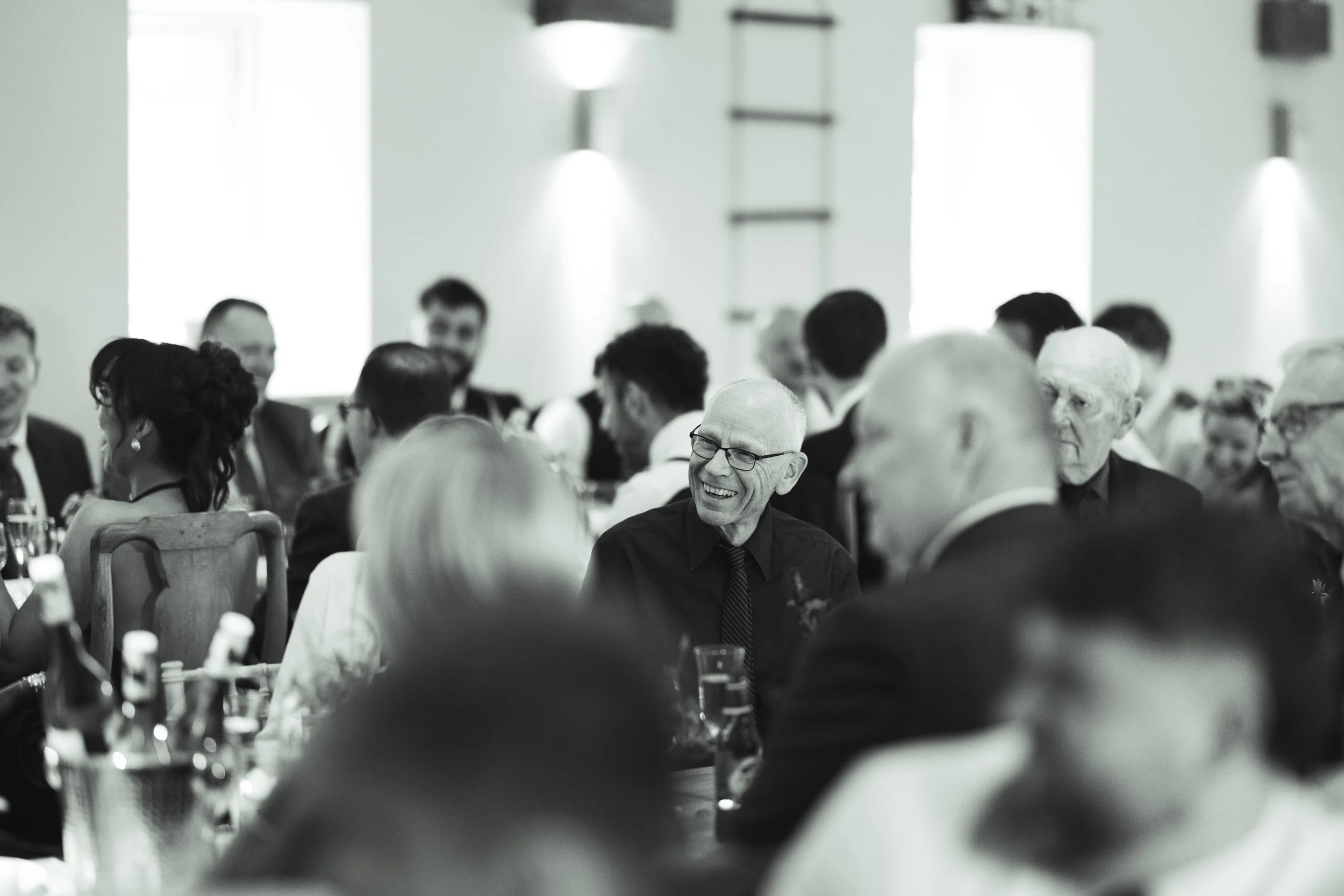 A group of people sitting at a long table during a social gathering or celebration, with one older man smiling and wearing glasses, in a well-lit room with large windows and minimal decorations.