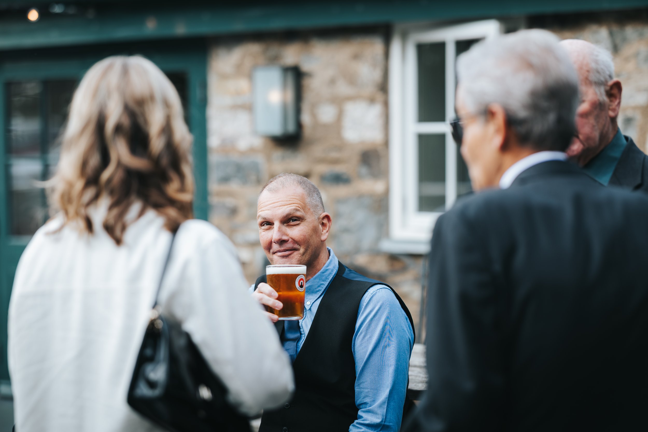 A man holding a glass of beer, engaging with three people at an outdoor gathering near a stone building.