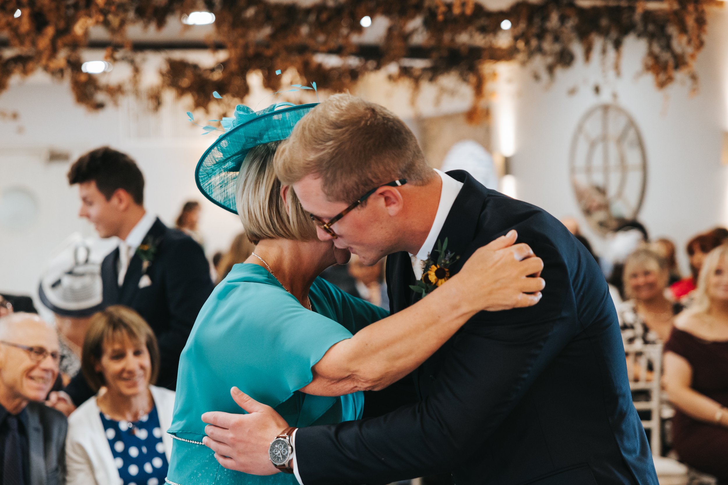 A young man and an older woman are sharing an emotional hug at a wedding, surrounded by other guests in the background.