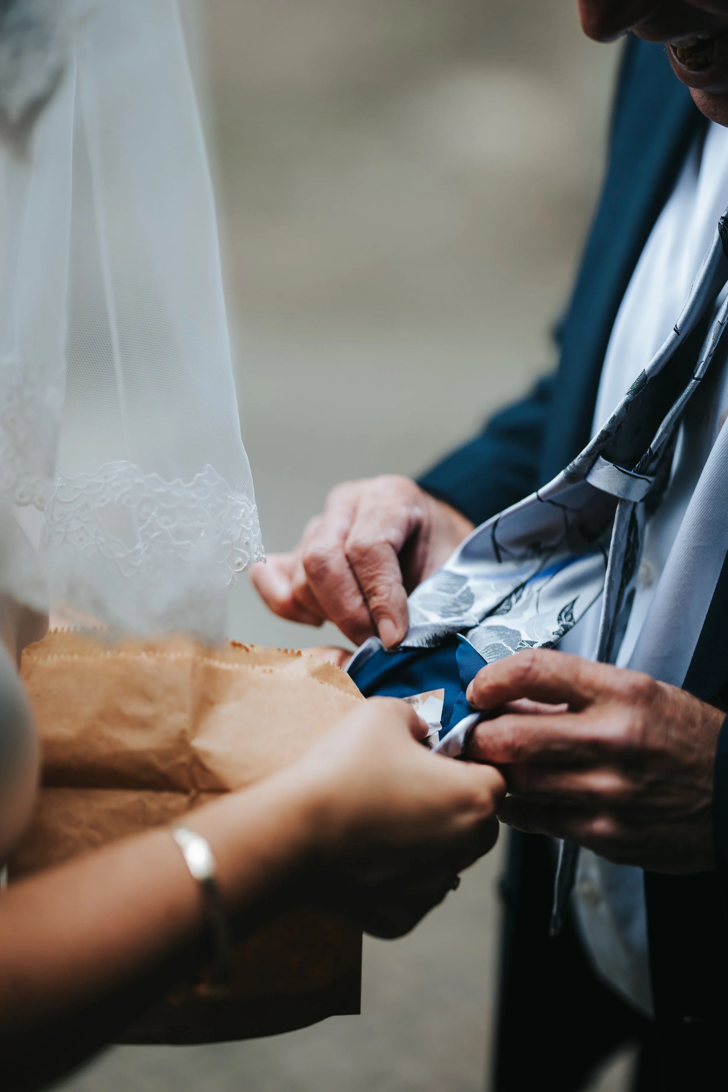 Close-up of a man in a suit with a patterned tie, adjusting his belt or waistband, with a woman holding a brown paper bag nearby during a wedding ceremony.