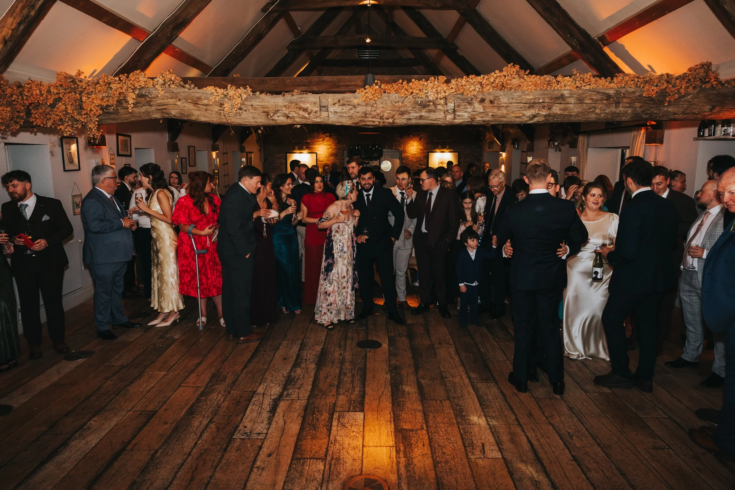 A large group of people dressed in formal attire gathered in a rustic-style indoor venue with wooden beams, celebrating at a wedding reception.