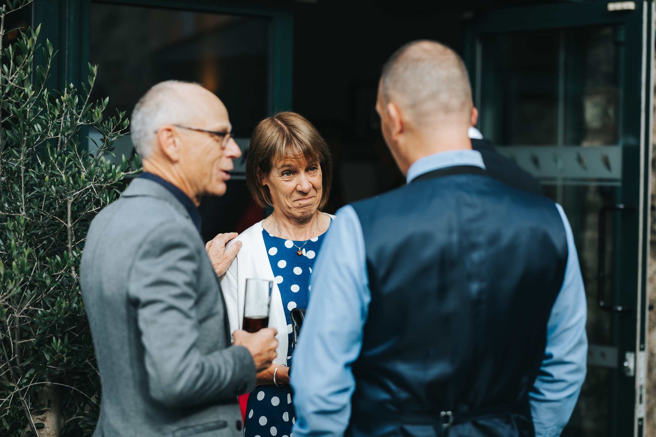 Three people at a social gathering, engaging in conversation outside. The woman in the center appears unsure or uncomfortable, holding a glass of wine, while the two men face her, one with gray hair and glasses, and the other with a shaved head, both