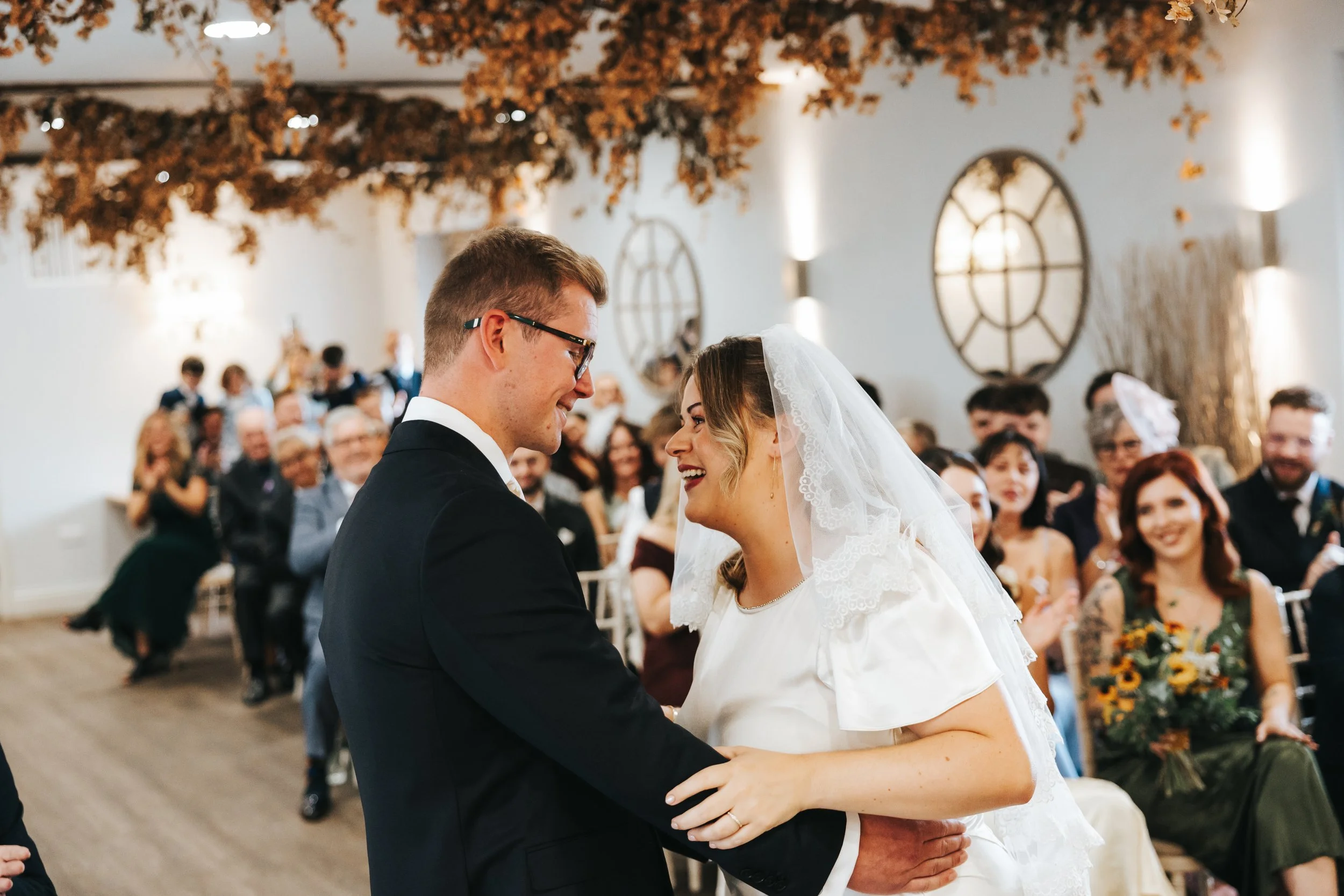 A bride and groom sharing a moment during their wedding ceremony, surrounded by friends and family in an indoor venue decorated with autumn leaves.