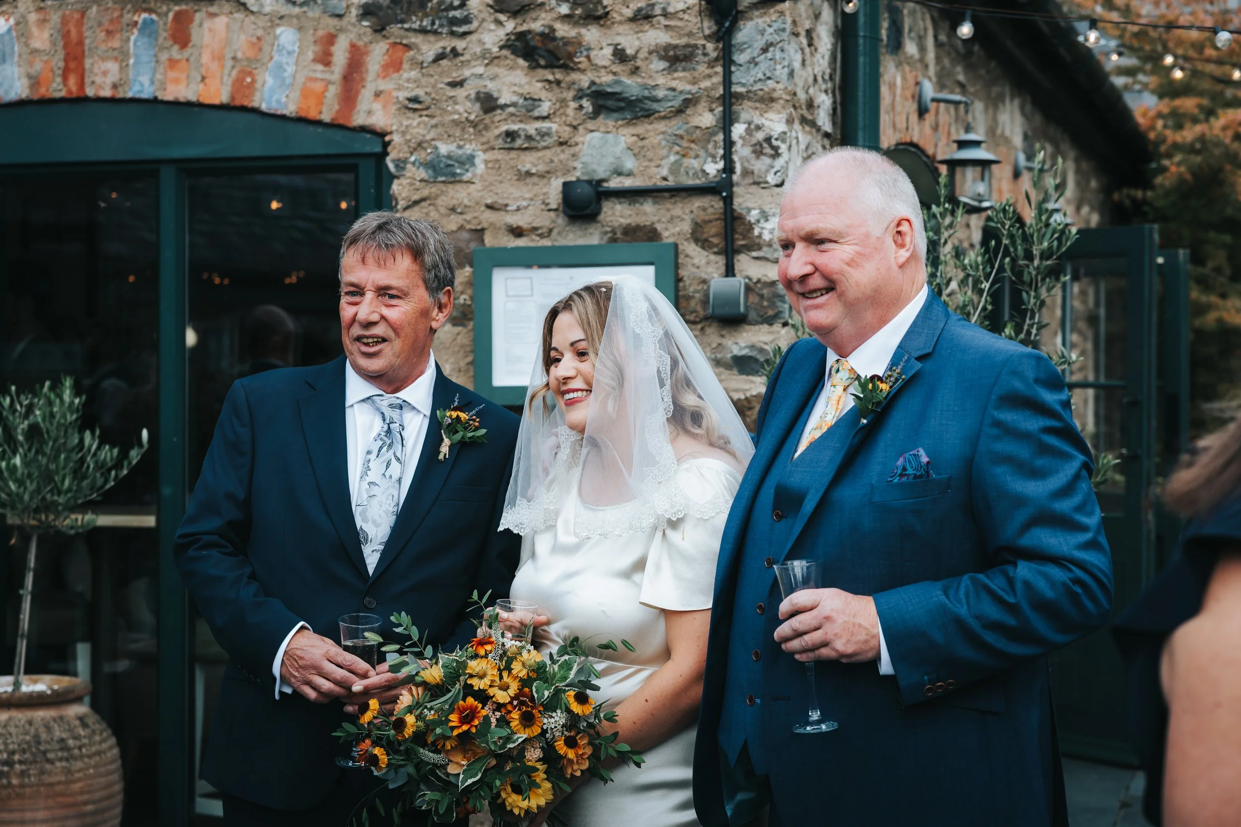 Bride and two men in suits smiling and holding drinks during a wedding celebration outside a stone building