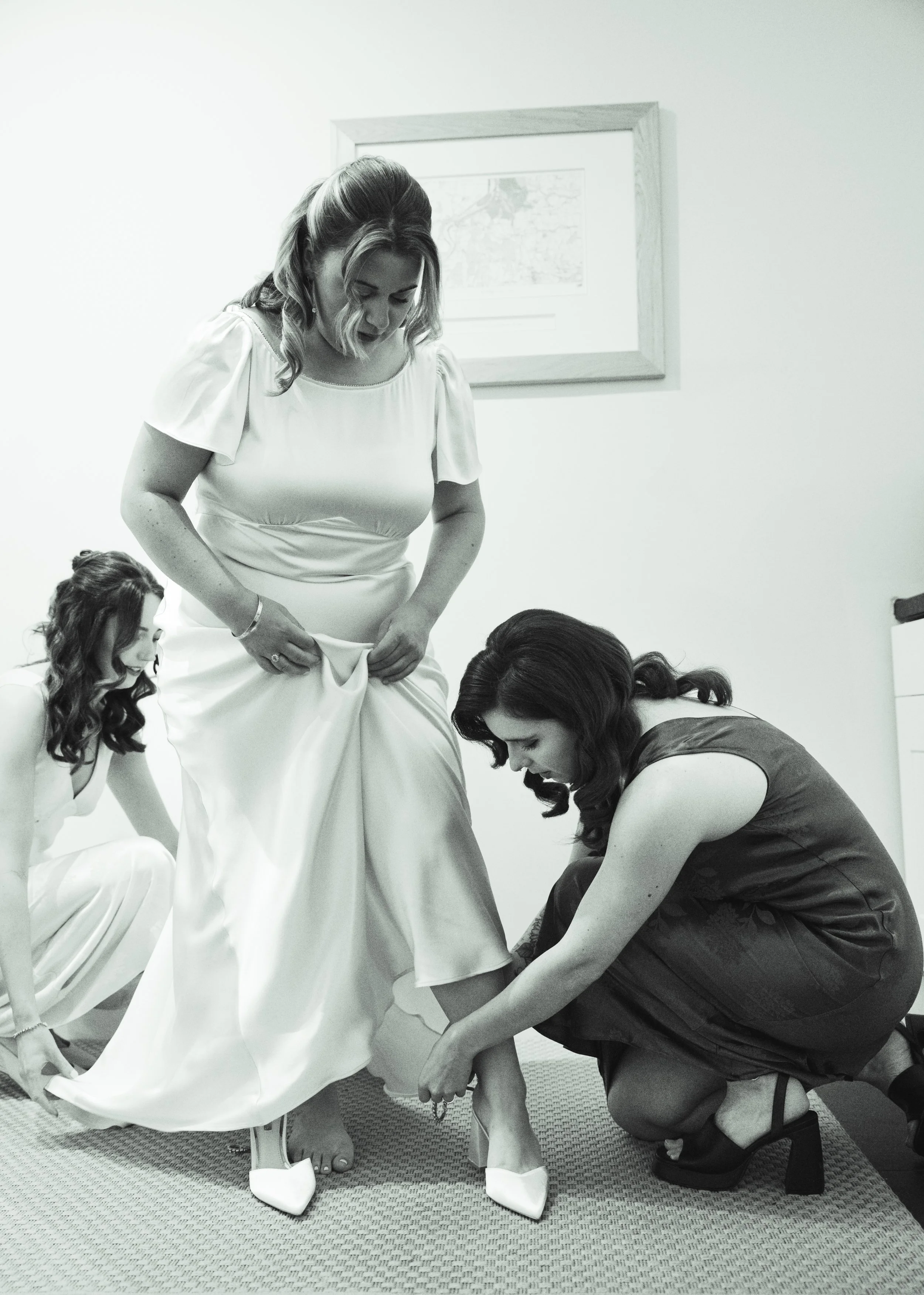 A bride getting her wedding shoes put on by a woman while another woman looks on in a room with a framed picture on the wall.