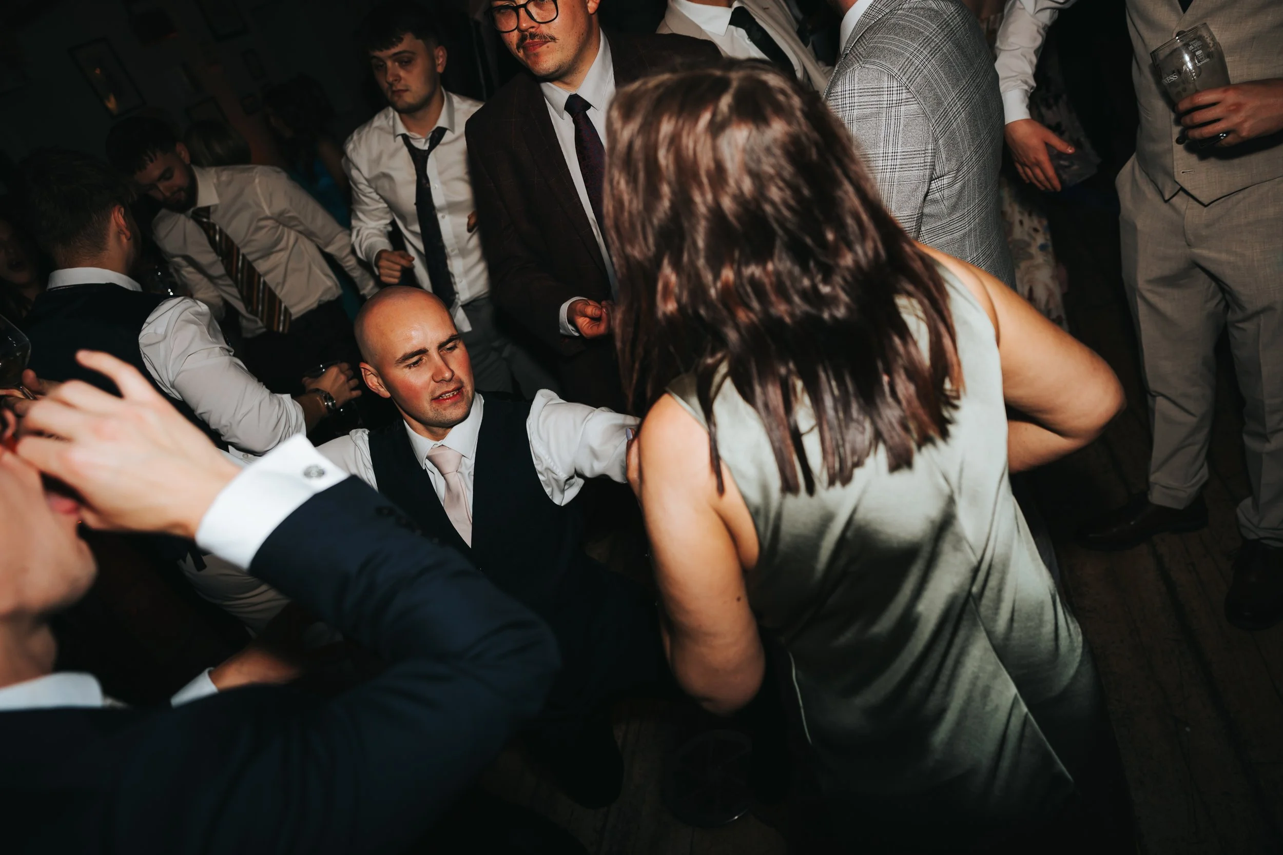 A group of people dancing at a party, with a man kneeling on the floor and a woman standing and facing him. The crowd is dressed in formal or semi-formal attire, and some are holding drinks.