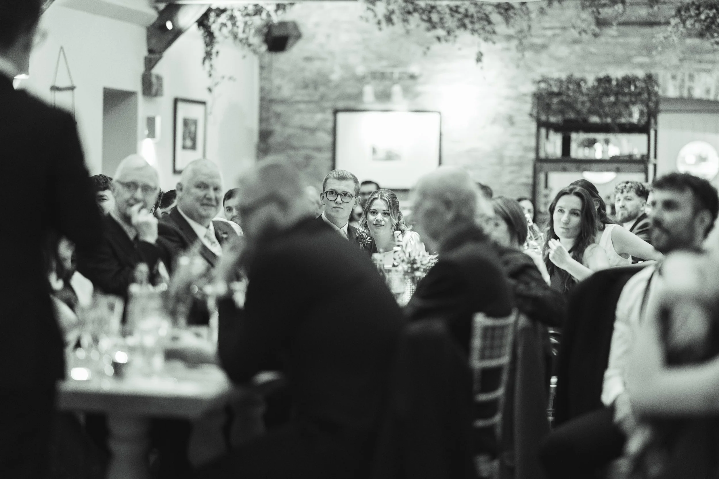People seated at a table during a formal event or gathering, attentively listening to a speaker in a cozy, rustic venue with brick walls and decorative plants.