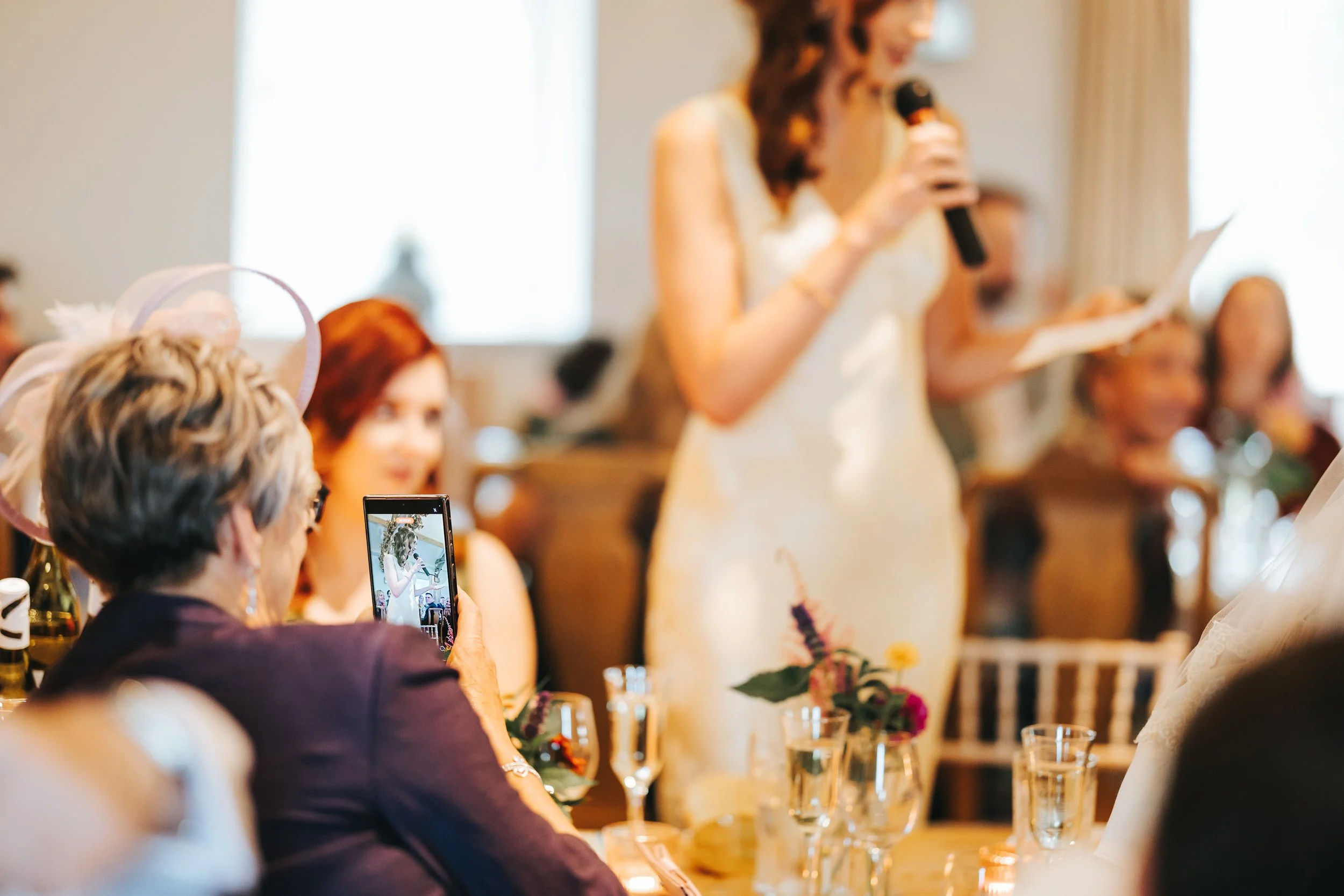 A woman in a white dress is giving a speech at a wedding reception, holding a microphone and reading from a paper. Guests, including an elderly woman with gray hair and a purple hat, are seated at a table with glasses and floral arrangements. One gue