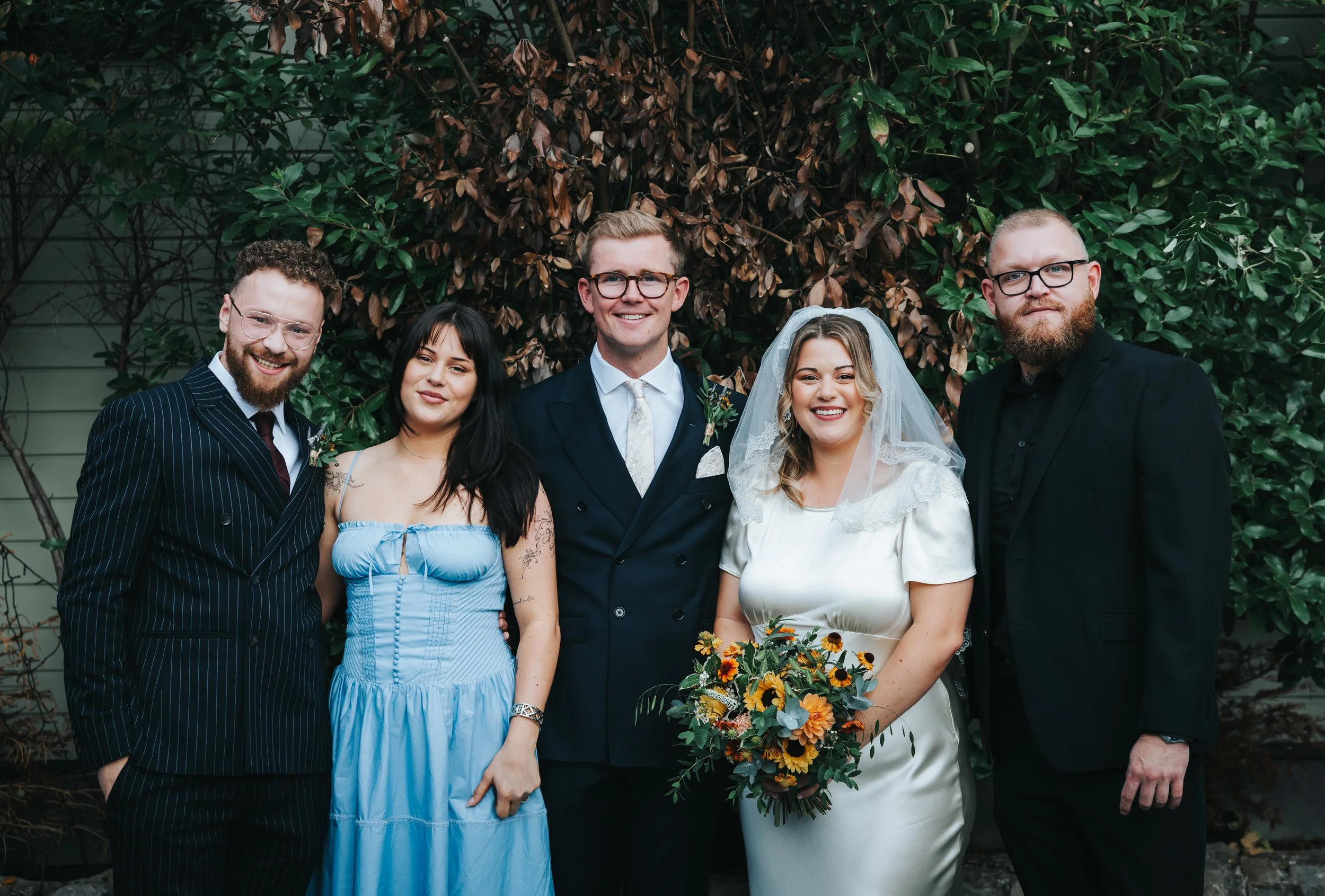 Group of six people at a wedding, three men and three women, standing outdoors in front of leafy foliage. The bride in white holding a bouquet, the groom in a dark suit, others in formal dresses and suits.