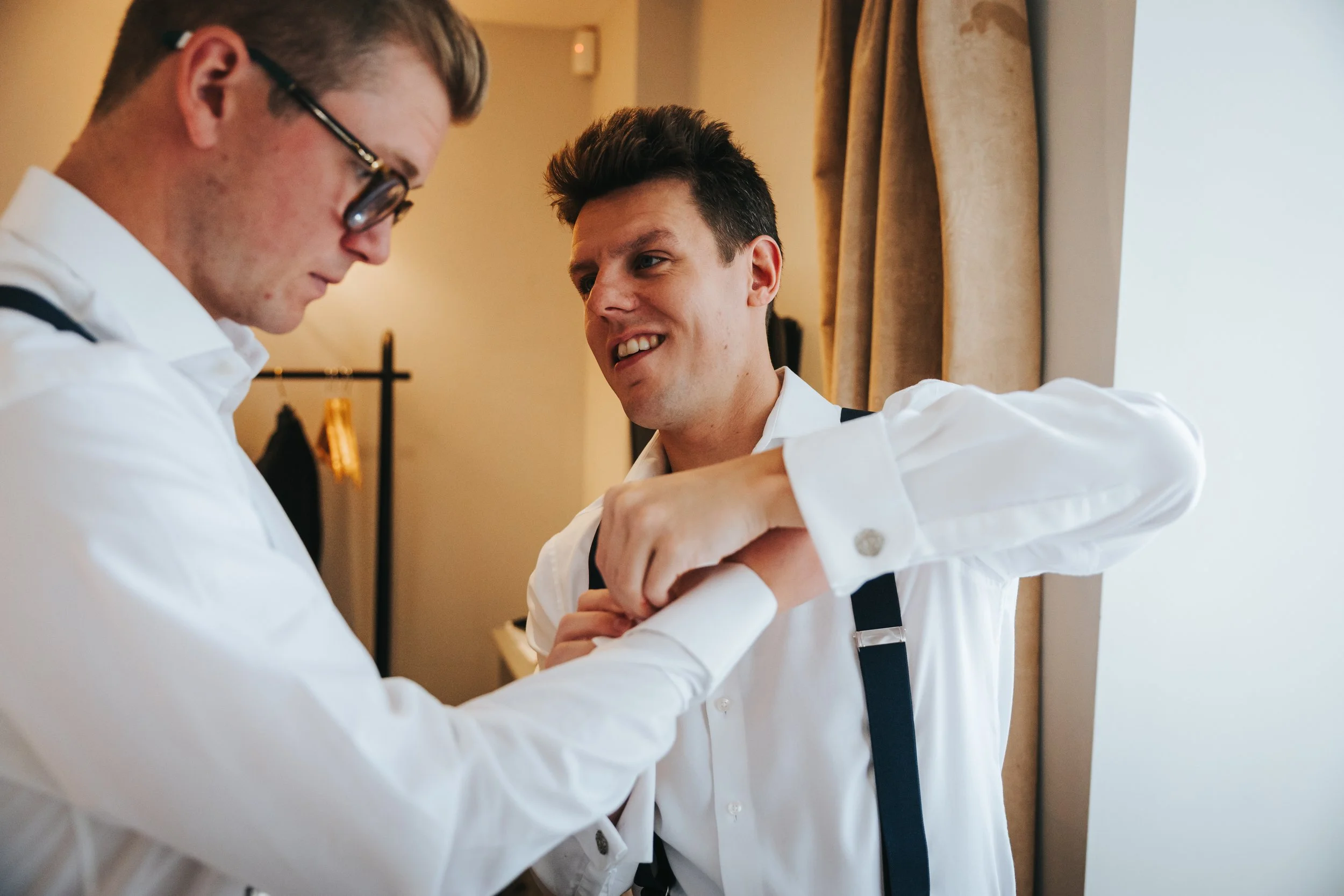 Two men in white shirts preparing for a formal event, one assisting the other with cufflinks, smiling and engaging in a friendly moment.