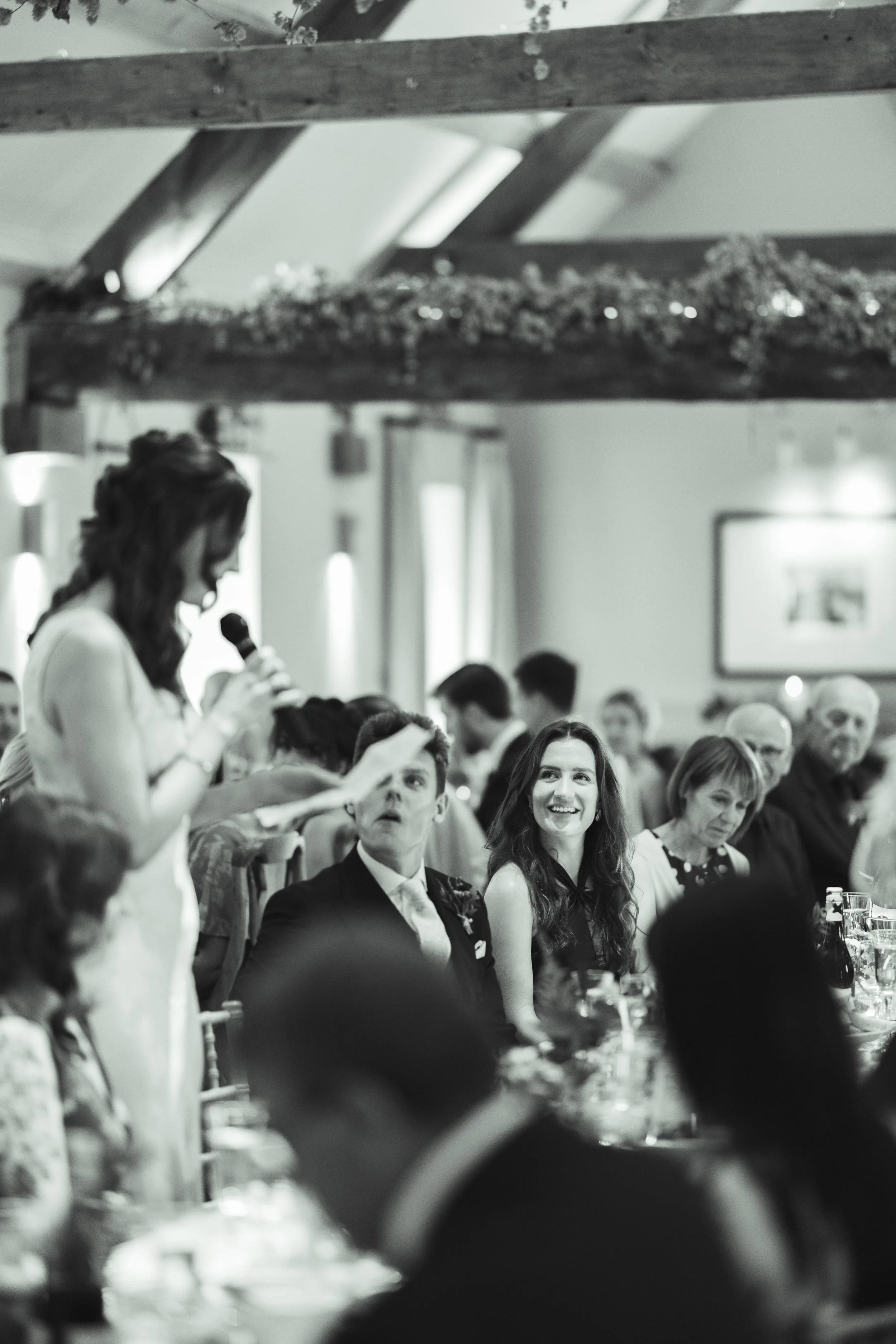 A woman is giving a speech at a formal event or celebration, with guests seated at tables watching her. The scene is in black and white.