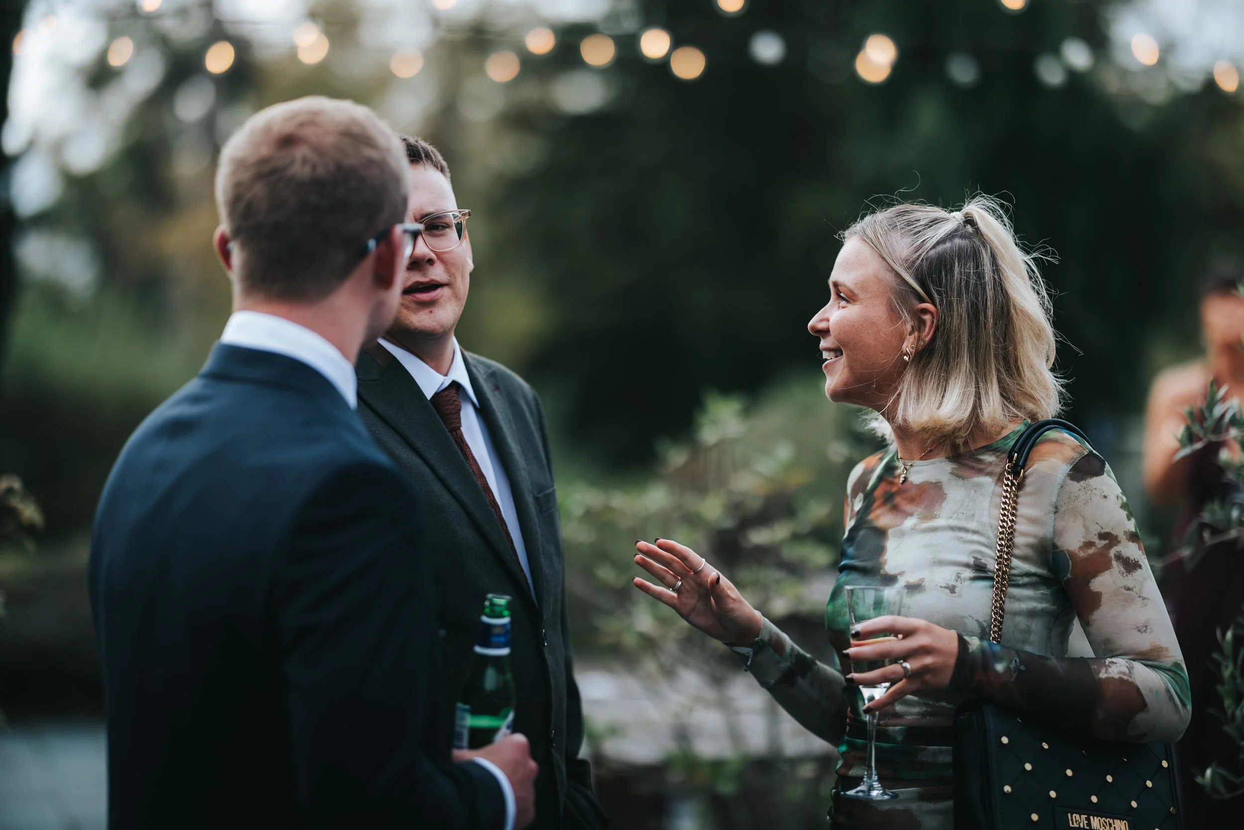 A group of three people, two men and one woman, are engaged in conversation outdoors during an evening event. The woman is holding a wine glass and smiling, while the men are holding drinks and listening attentively. The setting appears to be a garde