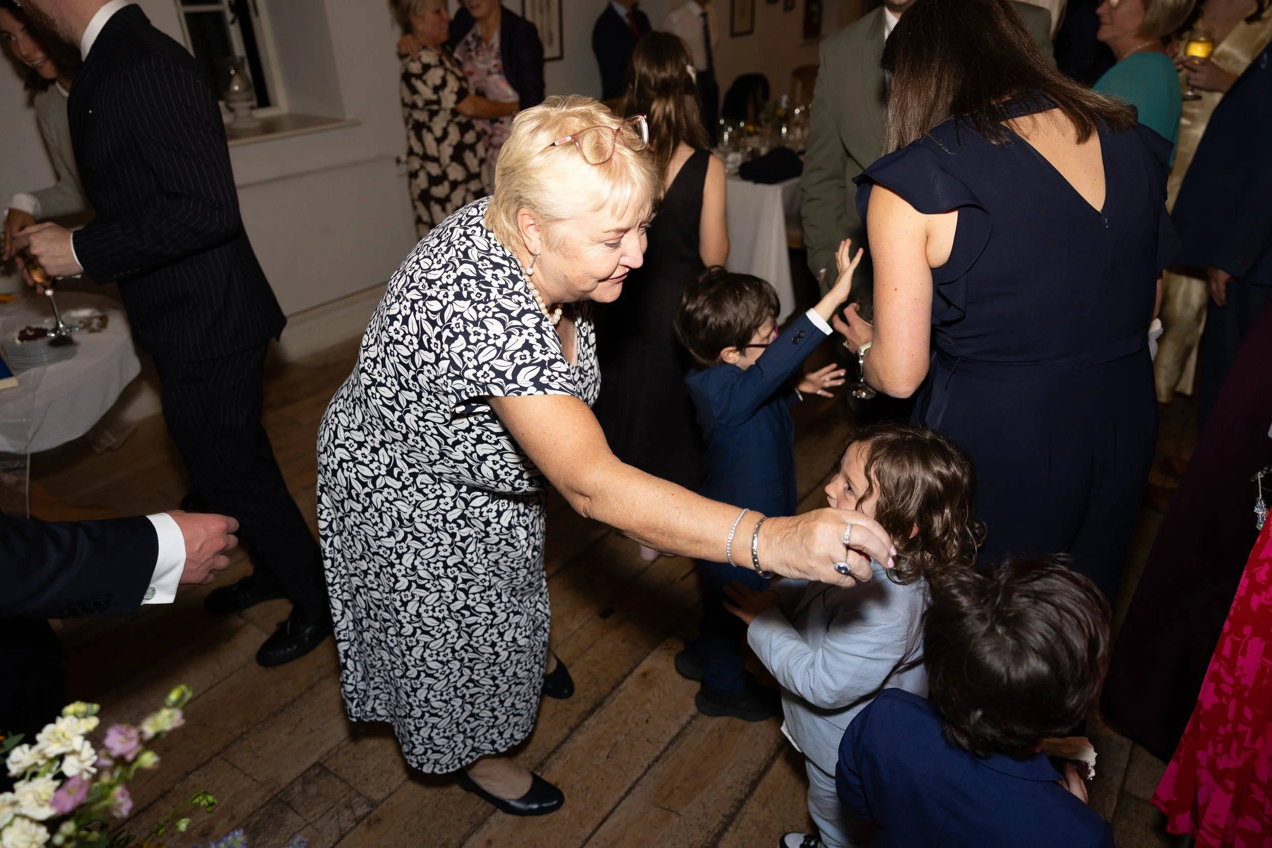 An elderly woman with glasses and pearl necklace dancing with a young girl at a lively social event, surrounded by other guests in formal attire.