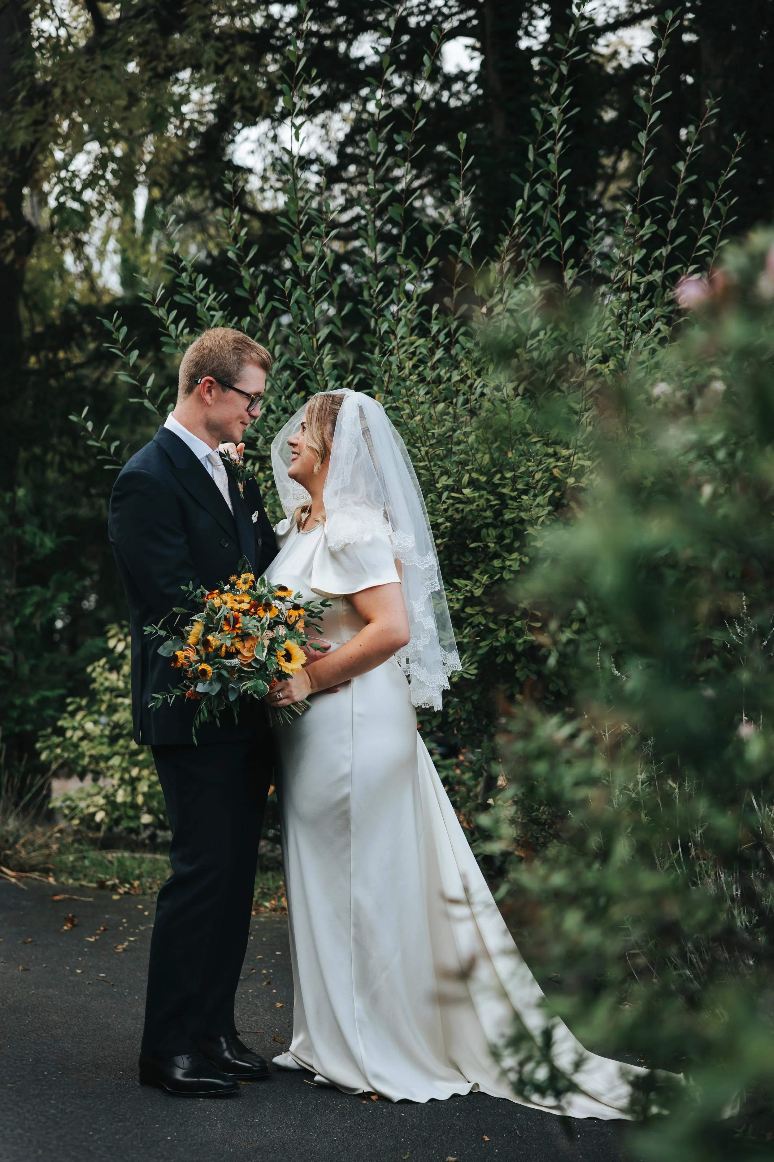 A bride and groom standing close together, smiling, with the bride holding a bouquet of flowers, surrounded by greenery during their wedding.