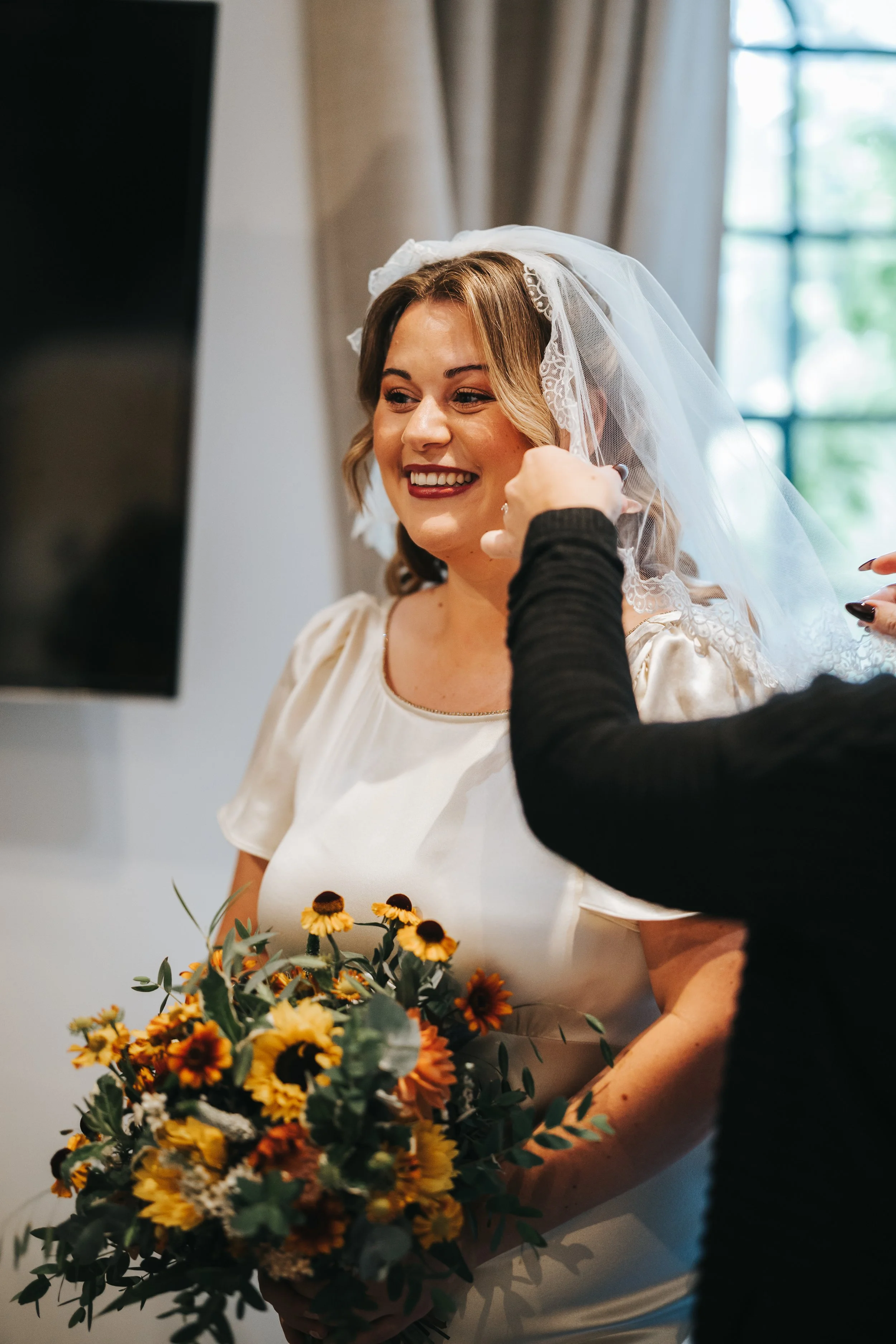 A bride with a bouquet of yellow and orange flowers, smiling as someone adjusts her veil.