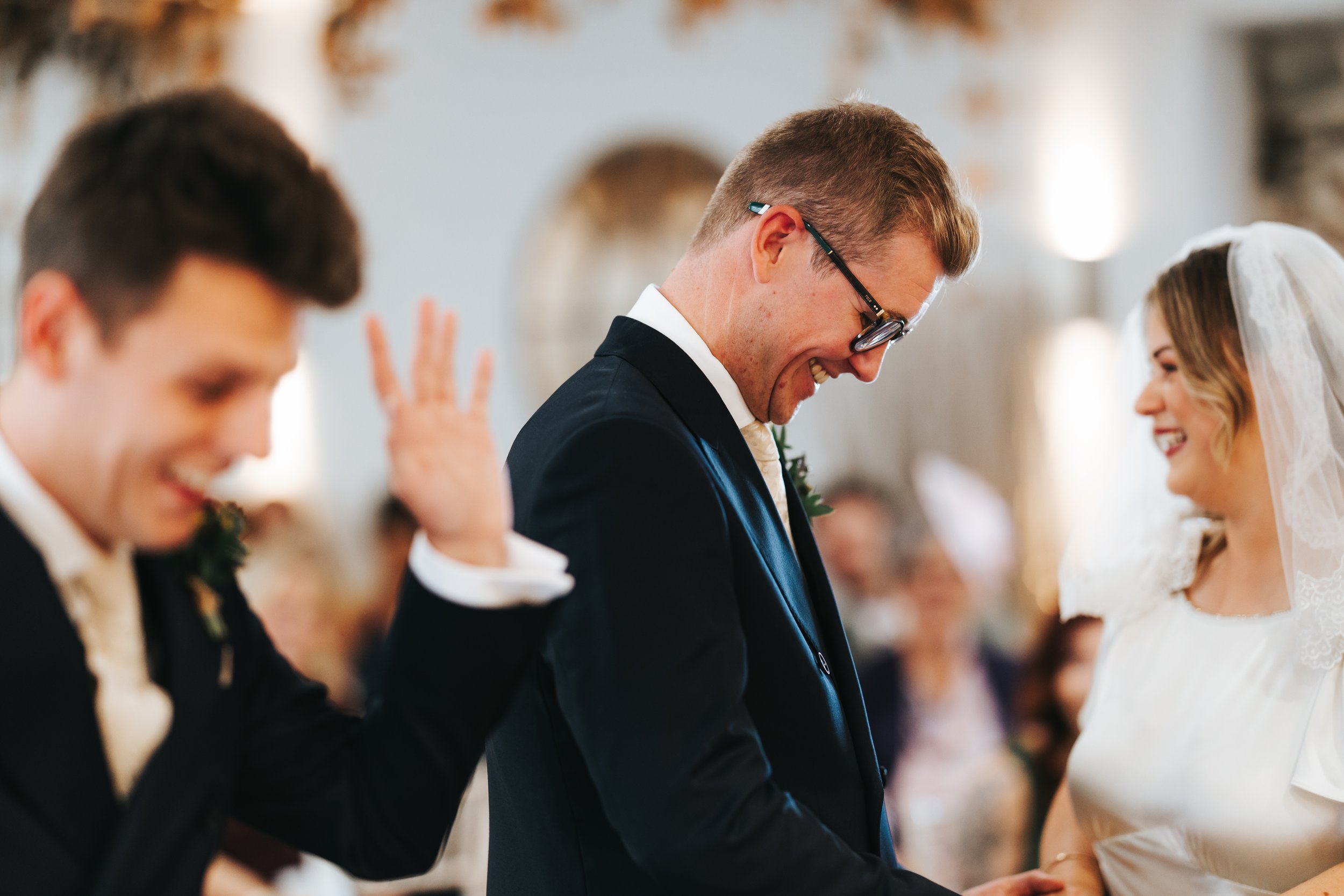 A joyful wedding ceremony with a groom, bride, and groomsman laughing and smiling inside a decorated venue.