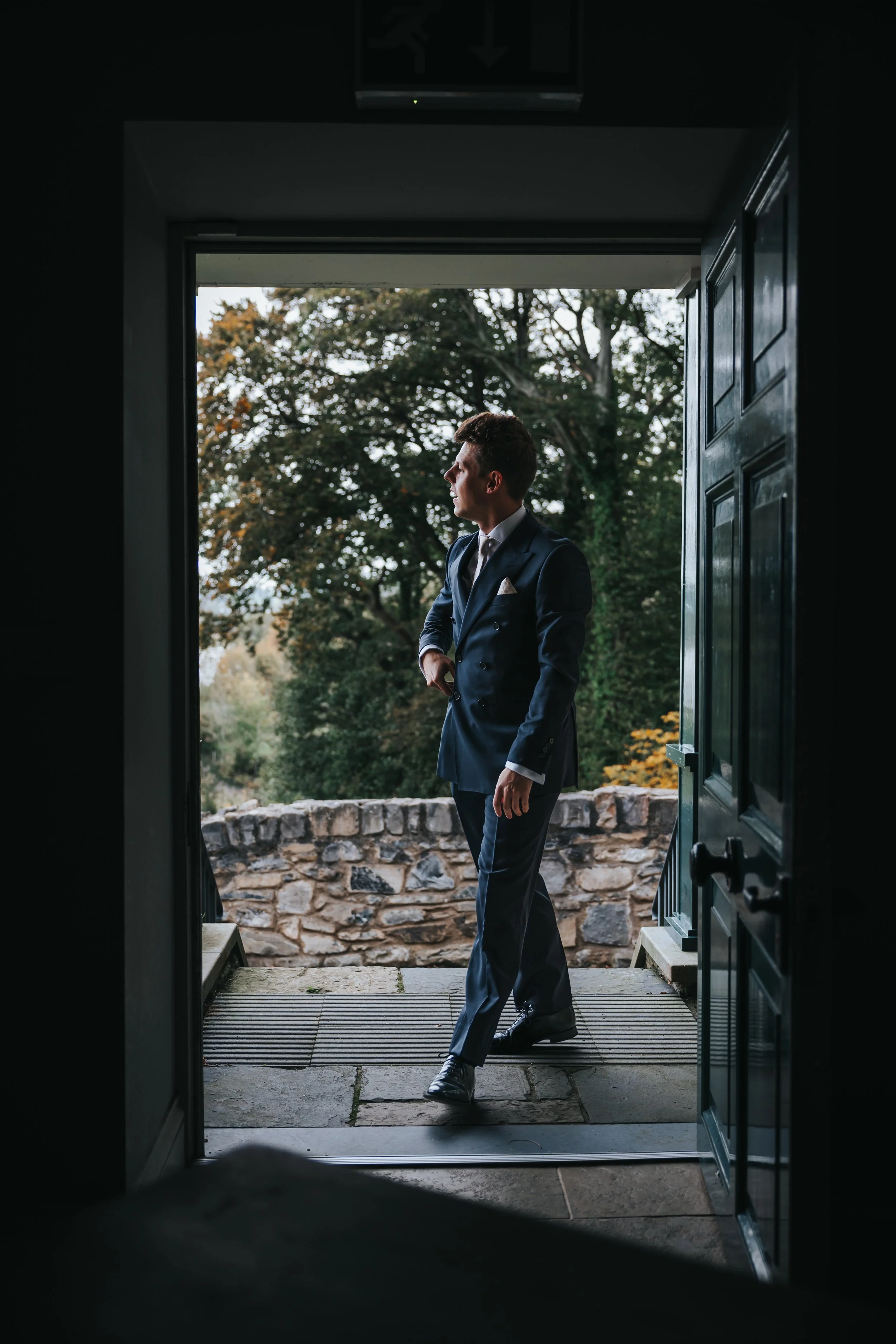 A man in a navy suit standing on a stone porch, looking to the side, outside through a door at a stone railing and trees.