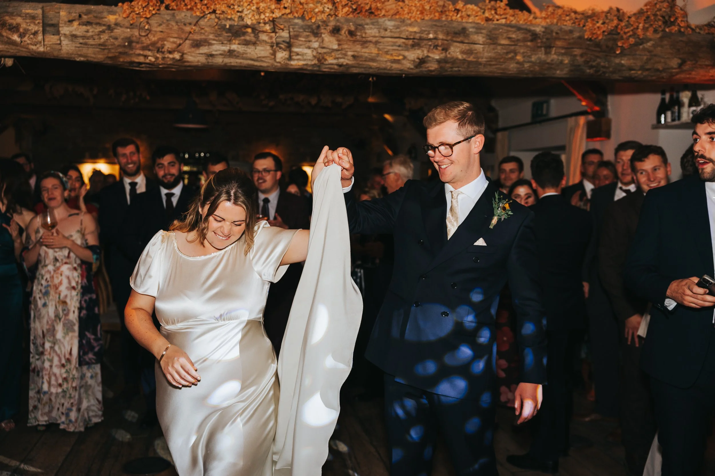 A bride and groom dancing at their wedding reception, surrounded by guests in formal attire, inside a rustic venue with wooden beams.