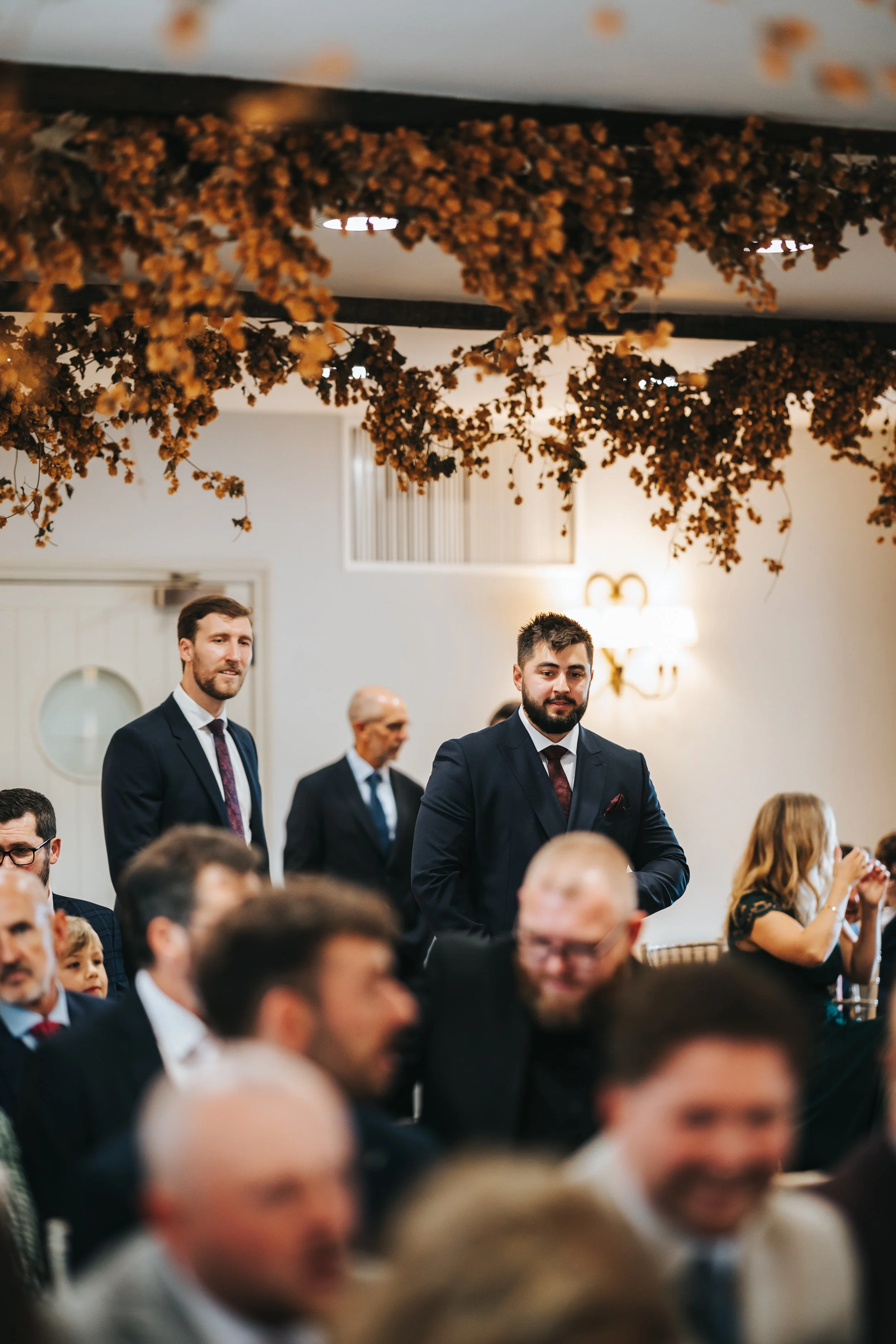 People in suits attending an event in a decorated indoor venue, with brown floral decorations hanging from the ceiling.