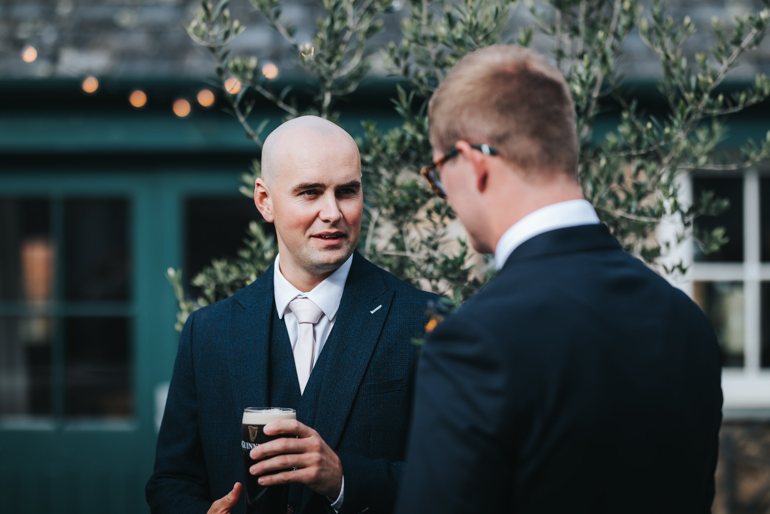 Two men in suits having a conversation outdoors, one holding a pint of Guinness stout.