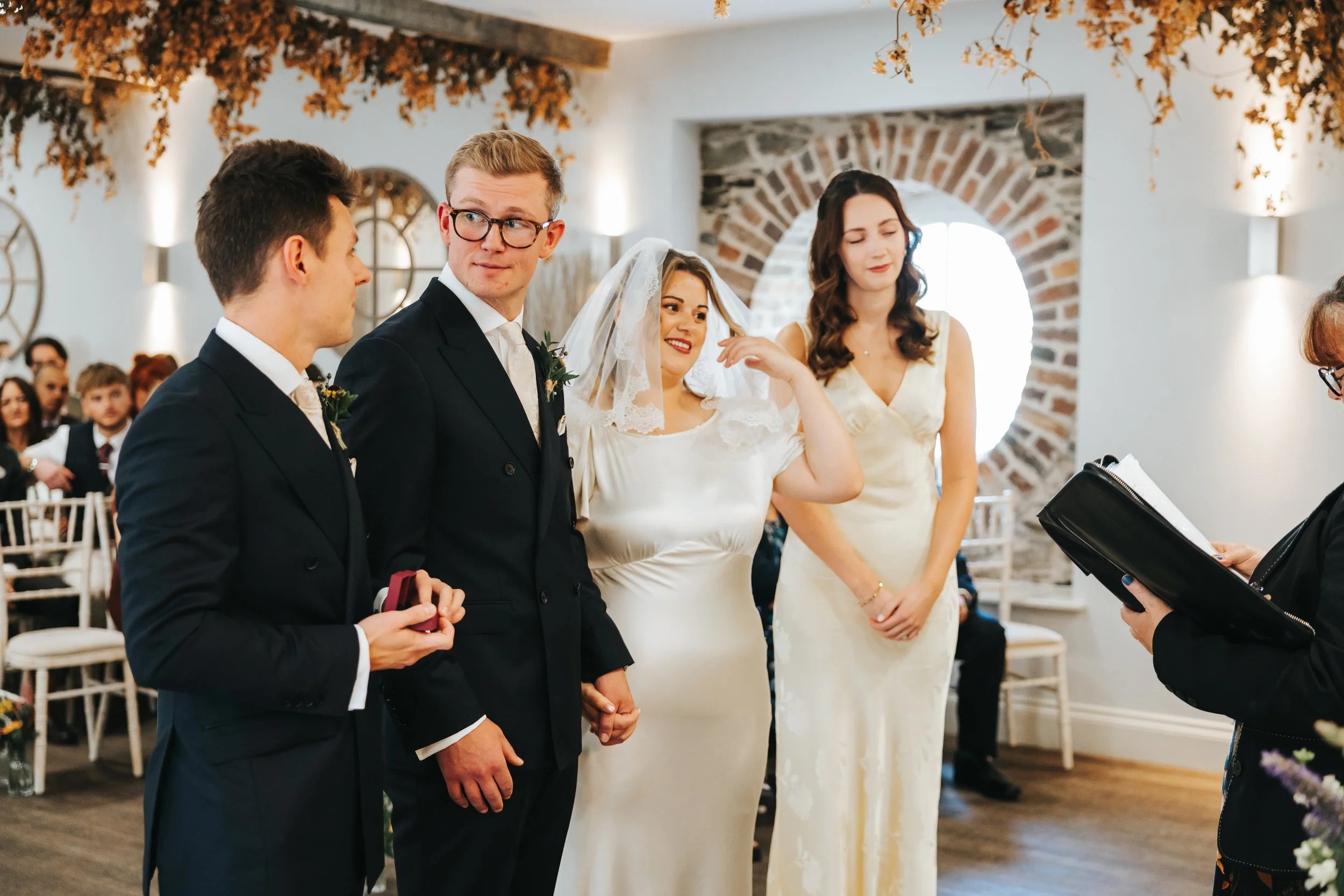 A wedding ceremony with a bride, groom, and two women standing in front of an officiant, inside a decorated venue with guests seated in the background.