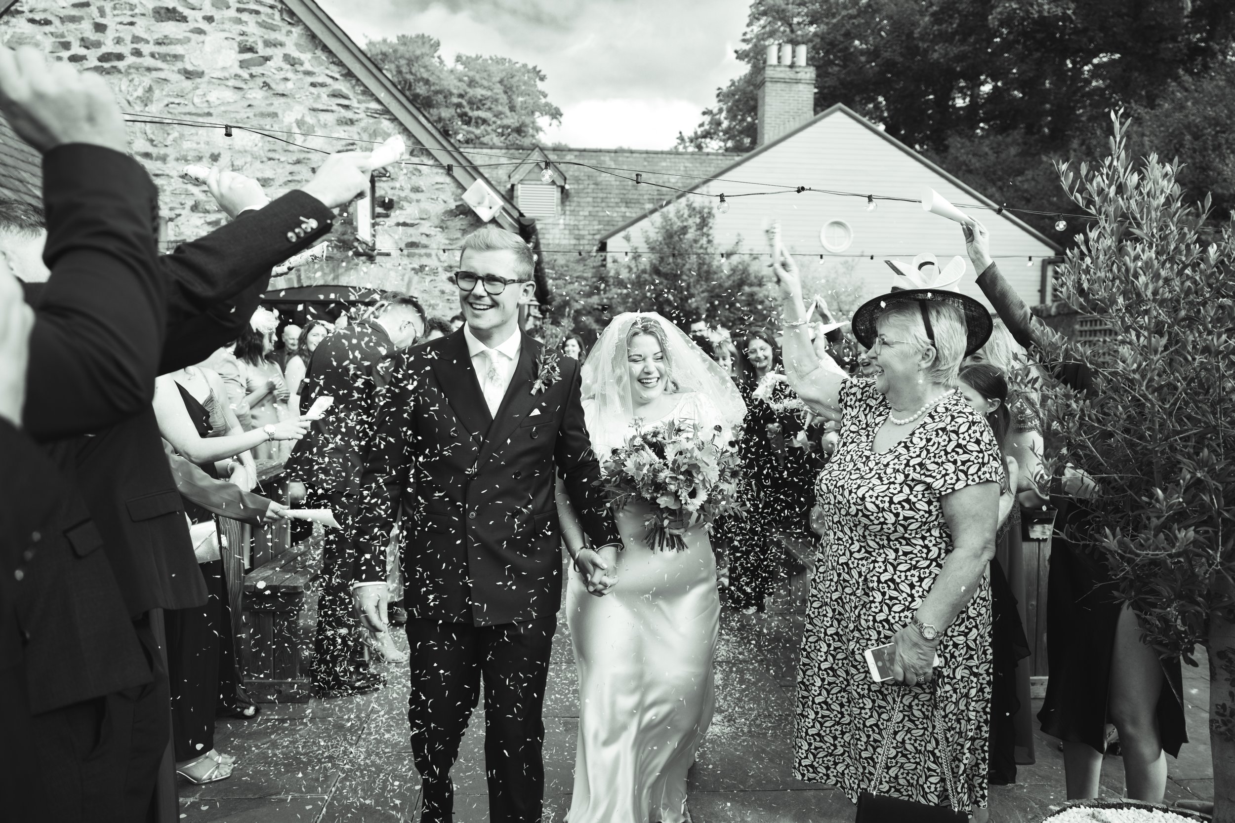 A bride and groom walking hand in hand through a crowd of friends and family at a wedding celebration, with people throwing confetti and celebrating.