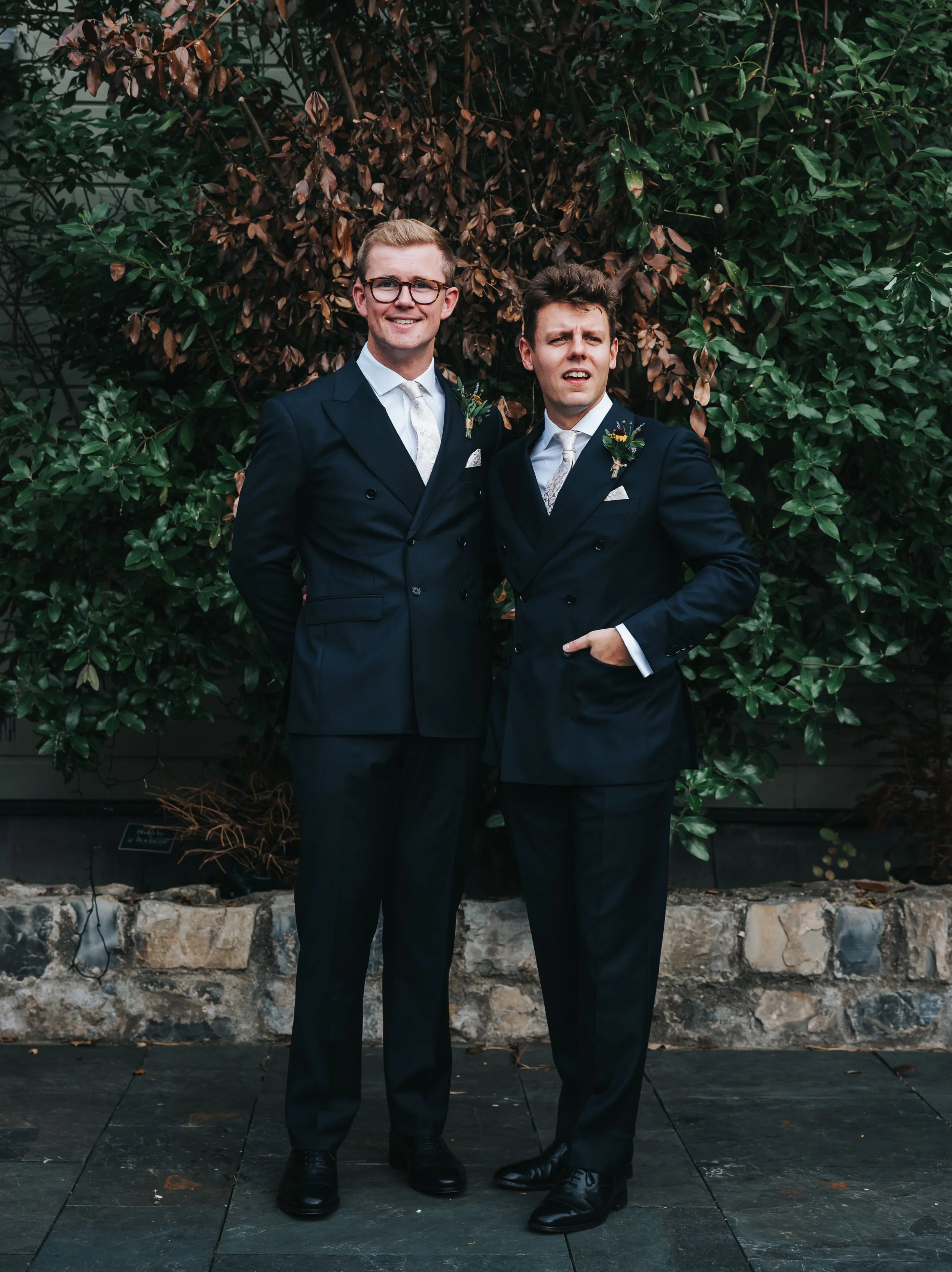 Two men in dark suits standing outdoors in front of lush green and brown foliage, smiling and making humorous facial expressions.