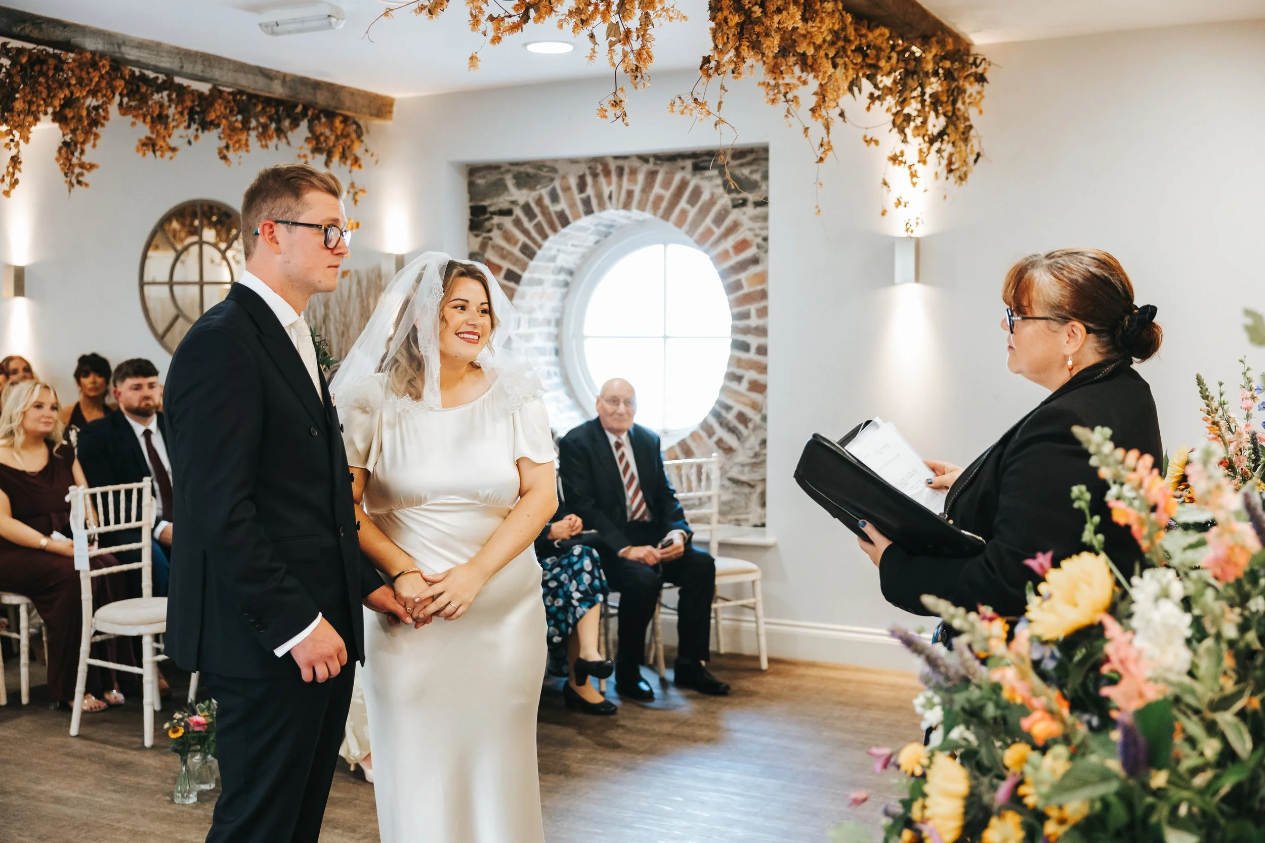 A couple stands during a wedding ceremony, holding hands and facing an officiant who is reading from a folder. The bride is wearing a white satin wedding dress and veil, and the groom is wearing a black suit with glasses. Guests are seated in the bac