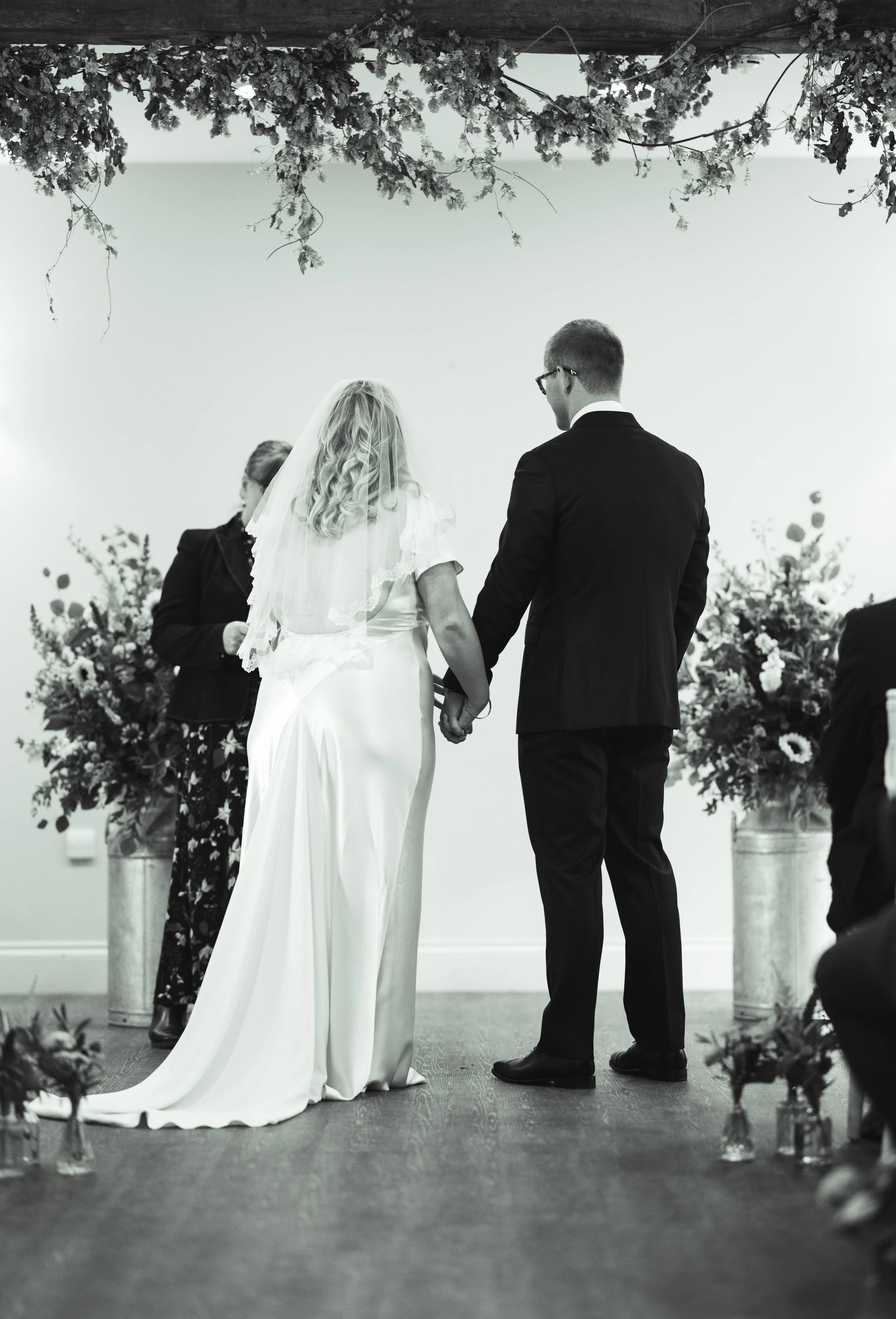 A couple getting married at a wedding ceremony, holding hands and facing an officiant, with floral arrangements on either side.
