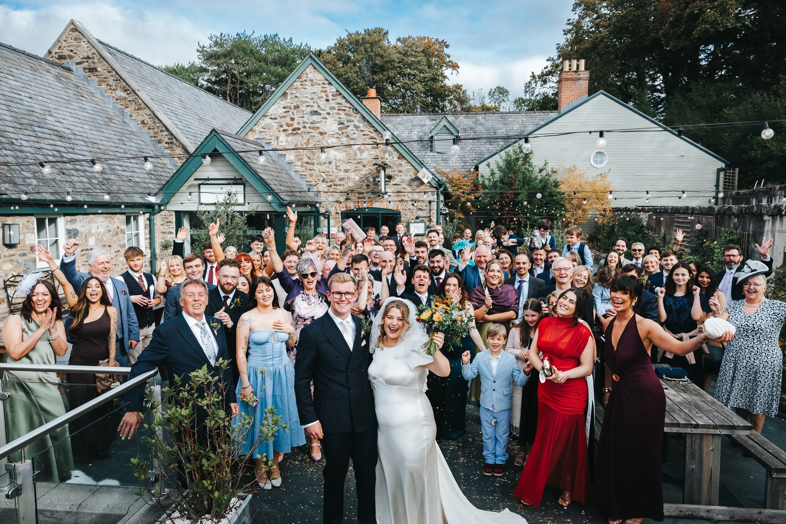 A large wedding celebration outdoors with the bride and groom in the front center, surrounded by friends and family on a patio with string lights and stone buildings in the background.