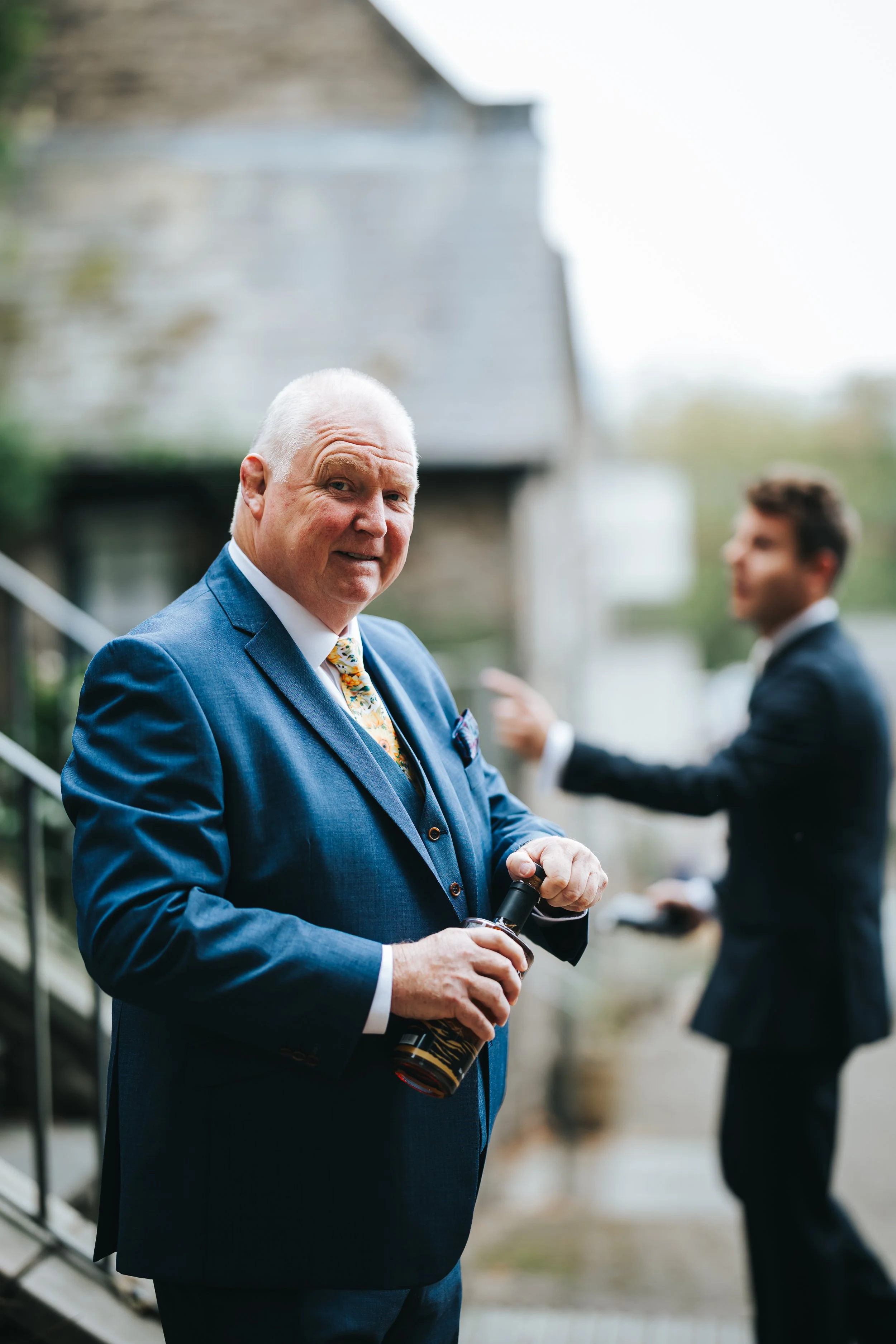 An older man in a blue suit holding a bottle of whiskey, standing outdoors during a daytime event. Another man in a tuxedo is blurred in the background, gesturing with his hand.