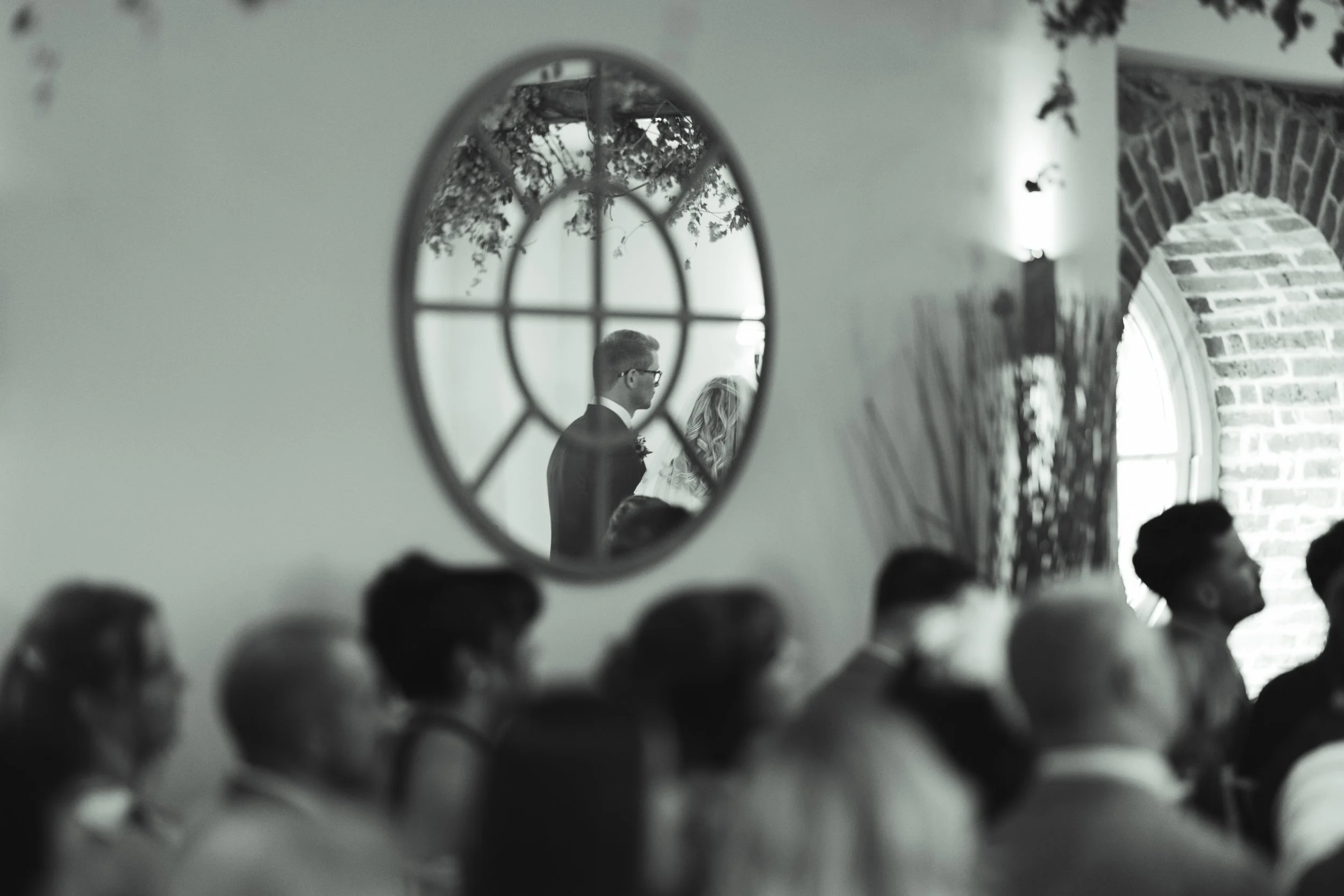 A black and white photo of a gathering with people watching an event, seen through an oval mirror on the wall that reflects a man and a woman, possibly at a wedding ceremony.
