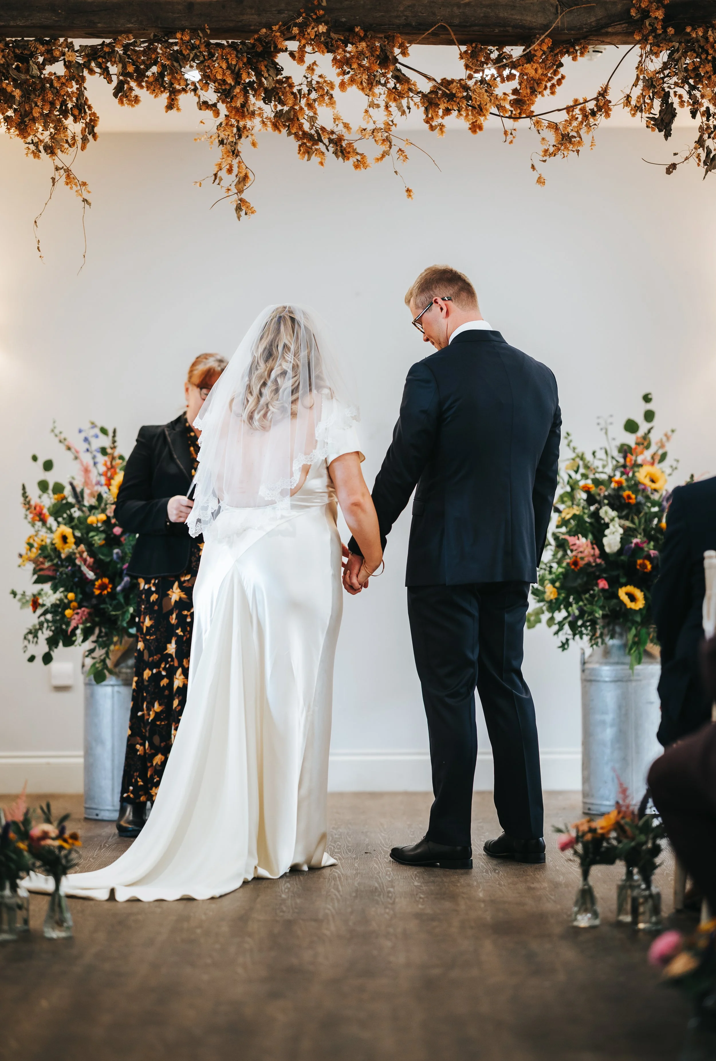 A bride and groom holding hands during a wedding ceremony, standing in front of a white wall decorated with large flower arrangements, with a woman officiating the ceremony.