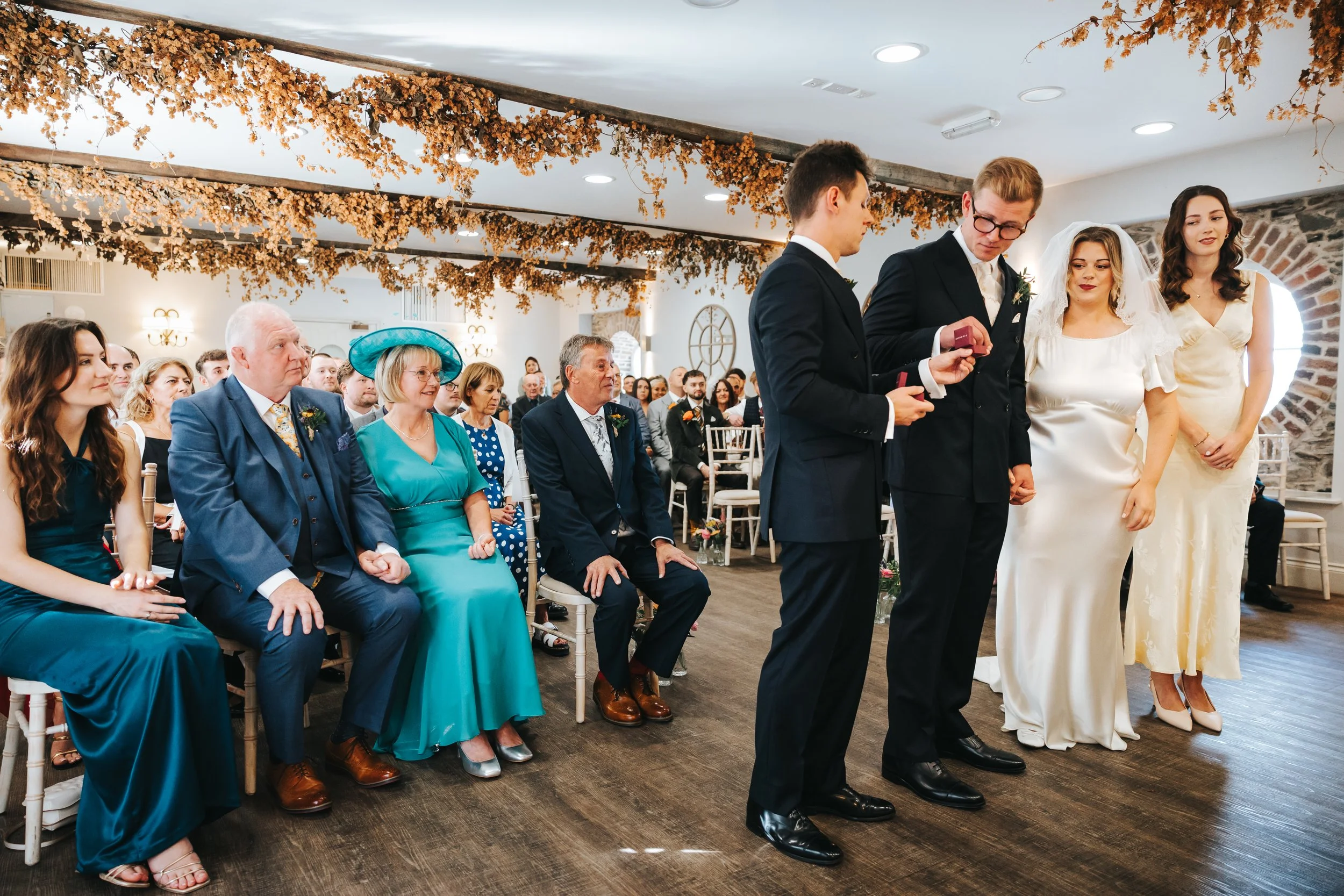 A wedding ceremony with guests seated, including older women in colorful dresses and men in suits. The bride and groom stand at the front, with the bride in a white wedding dress and veil, and the groom in a black suit. The venue is decorated with ha