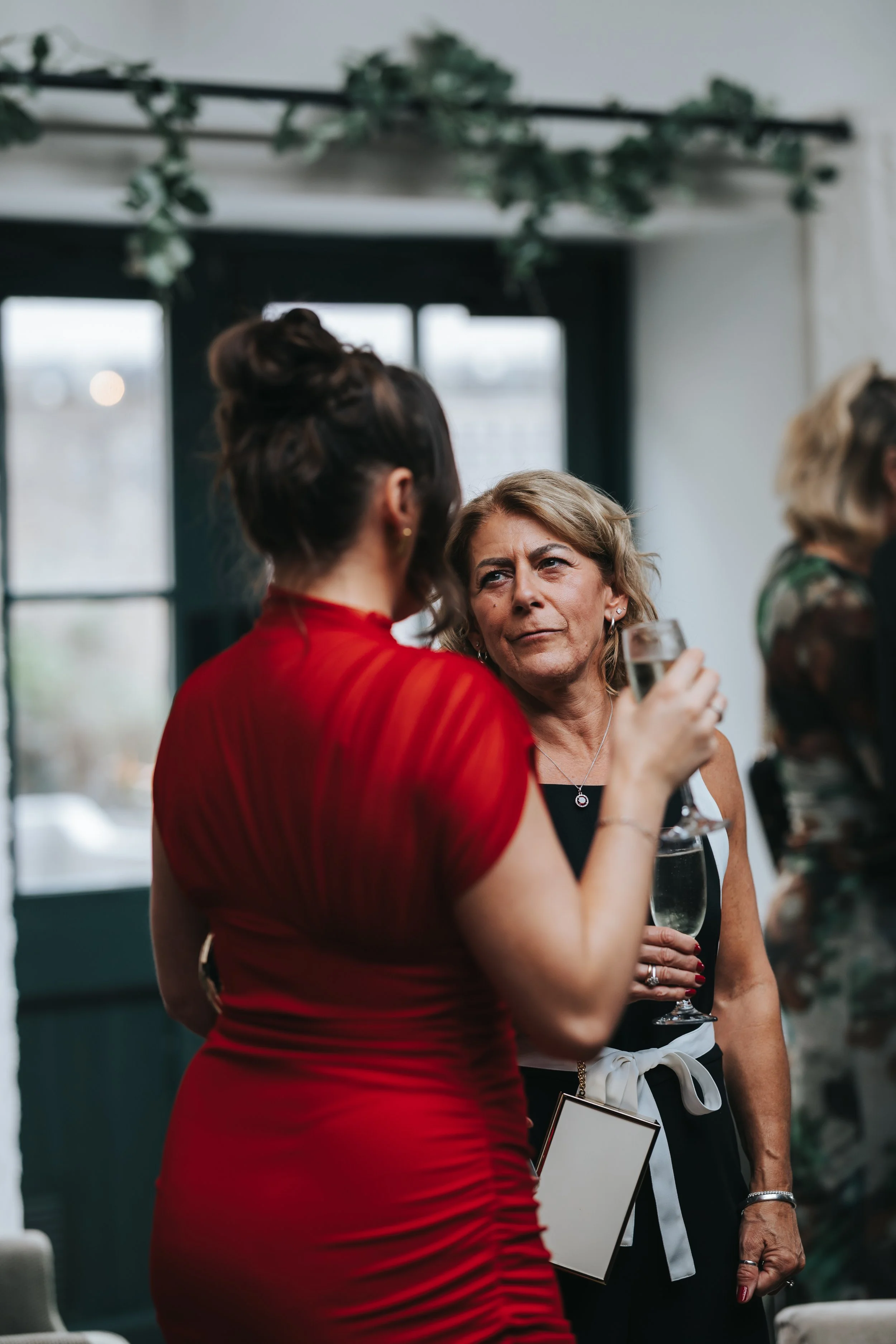 Two women having a conversation at a social gathering, one in a red dress holding a champagne glass, the other in a black dress with a white ribbon around her waist, also with a glass of champagne.