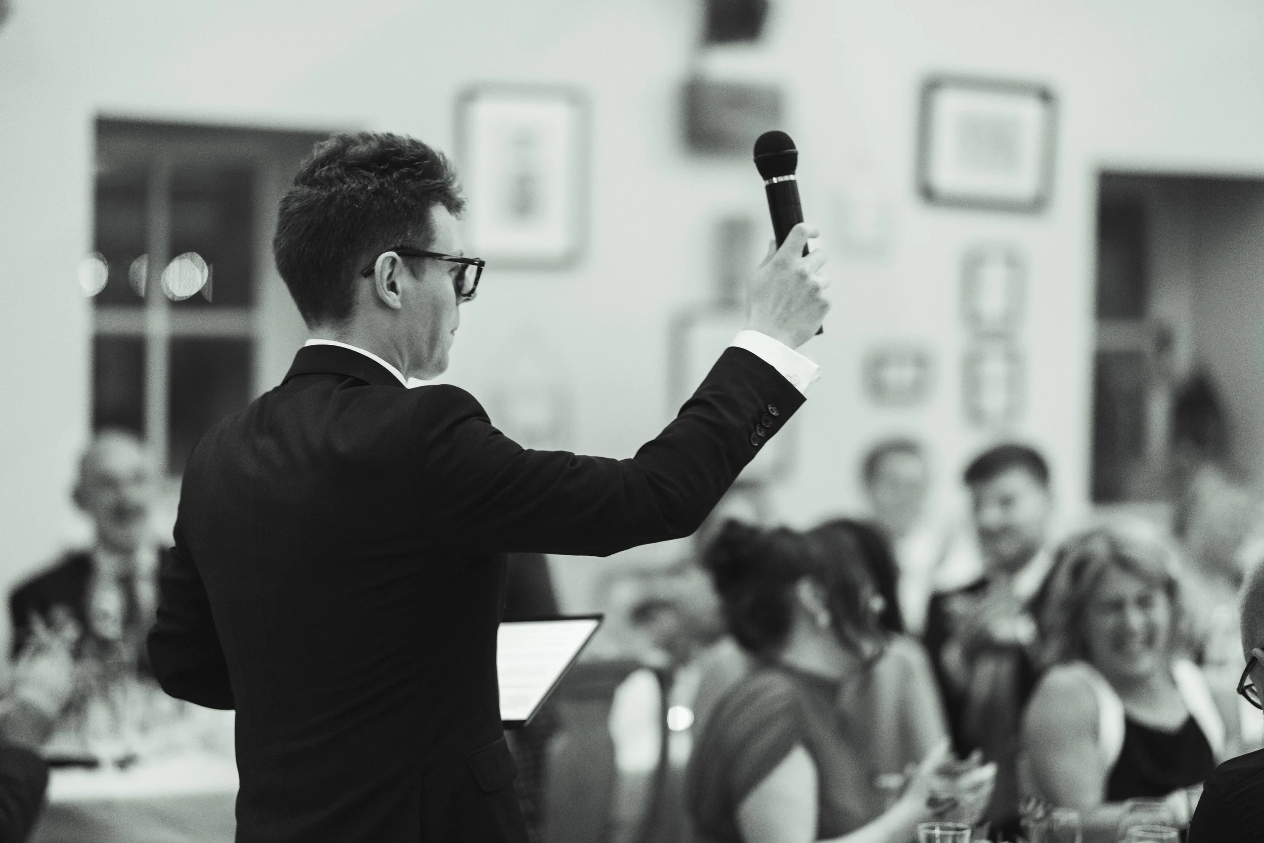 Man in suit giving speech or presentation at a formal event or meeting, holding a microphone, with an audience in the background.
