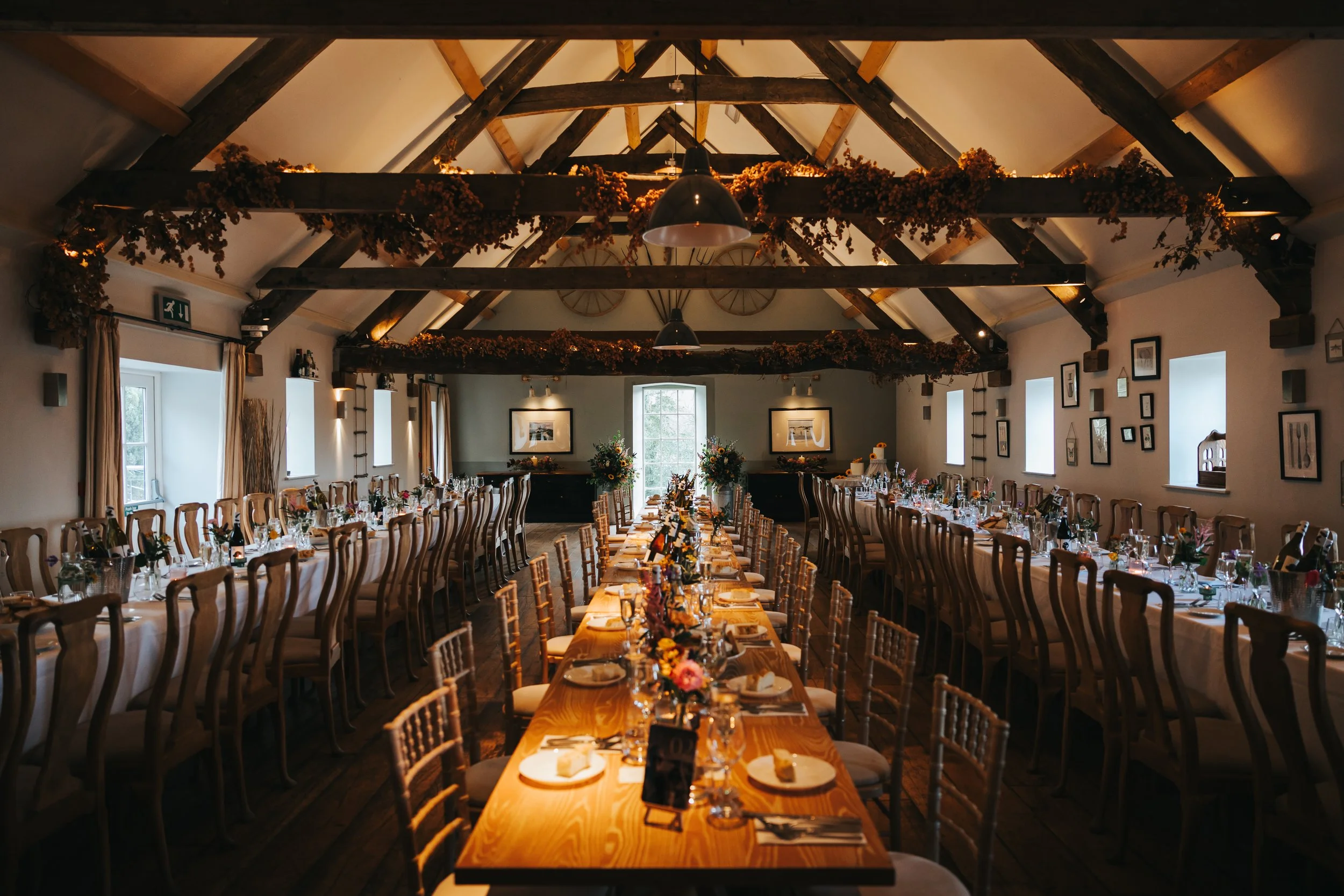 Elegant banquet hall decorated for a wedding reception, with long tables set with plates, glasses, and floral centerpieces, and wooden chairs arranged on both sides under a vaulted ceiling with exposed beams.